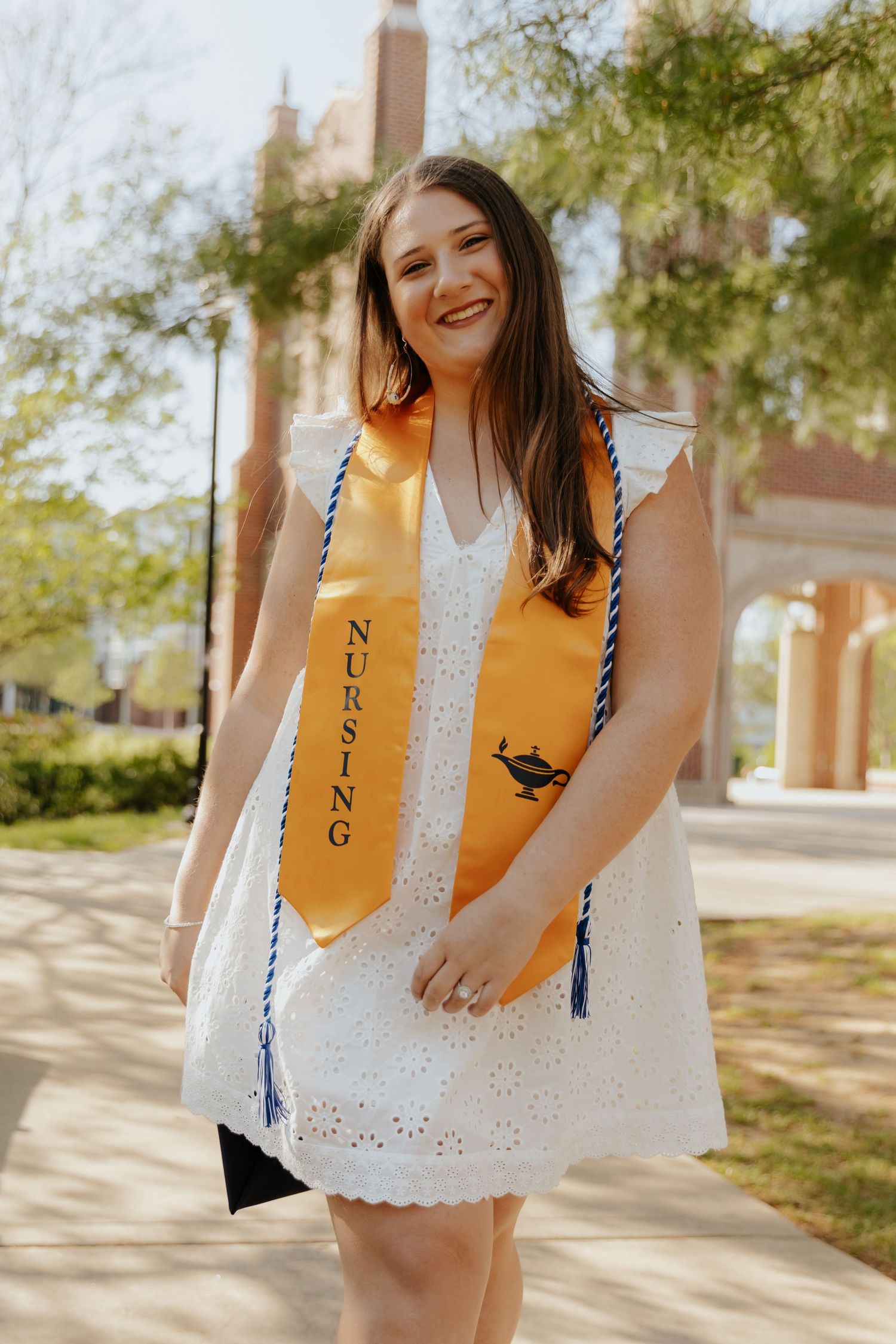 A graduate in a white eyelet dress wearing a yellow nursing graduation stole with blue and white cords smiles outdoors.