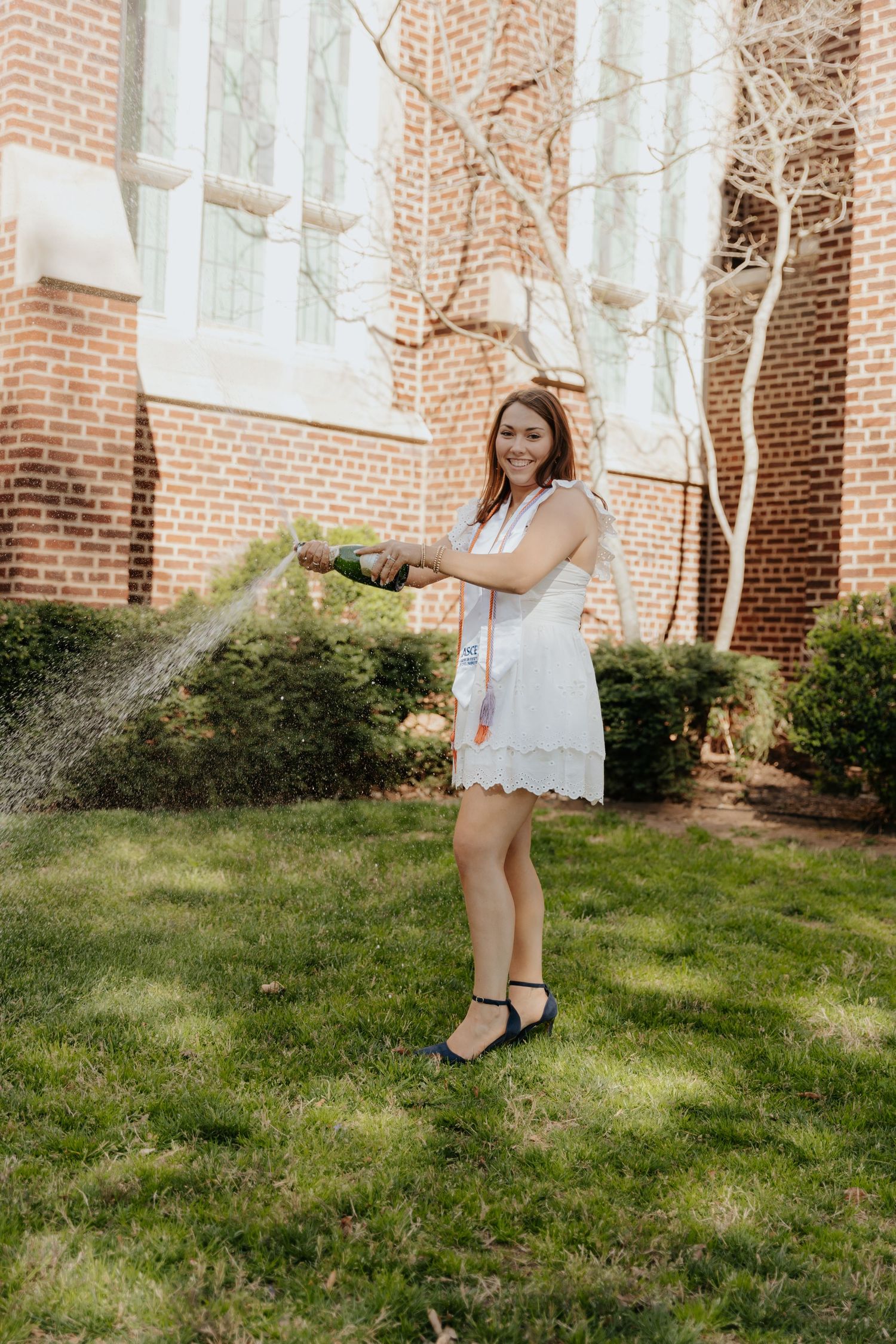 A person in a white summer dress poses outdoors while watering plants on a lawn next to a brick building.