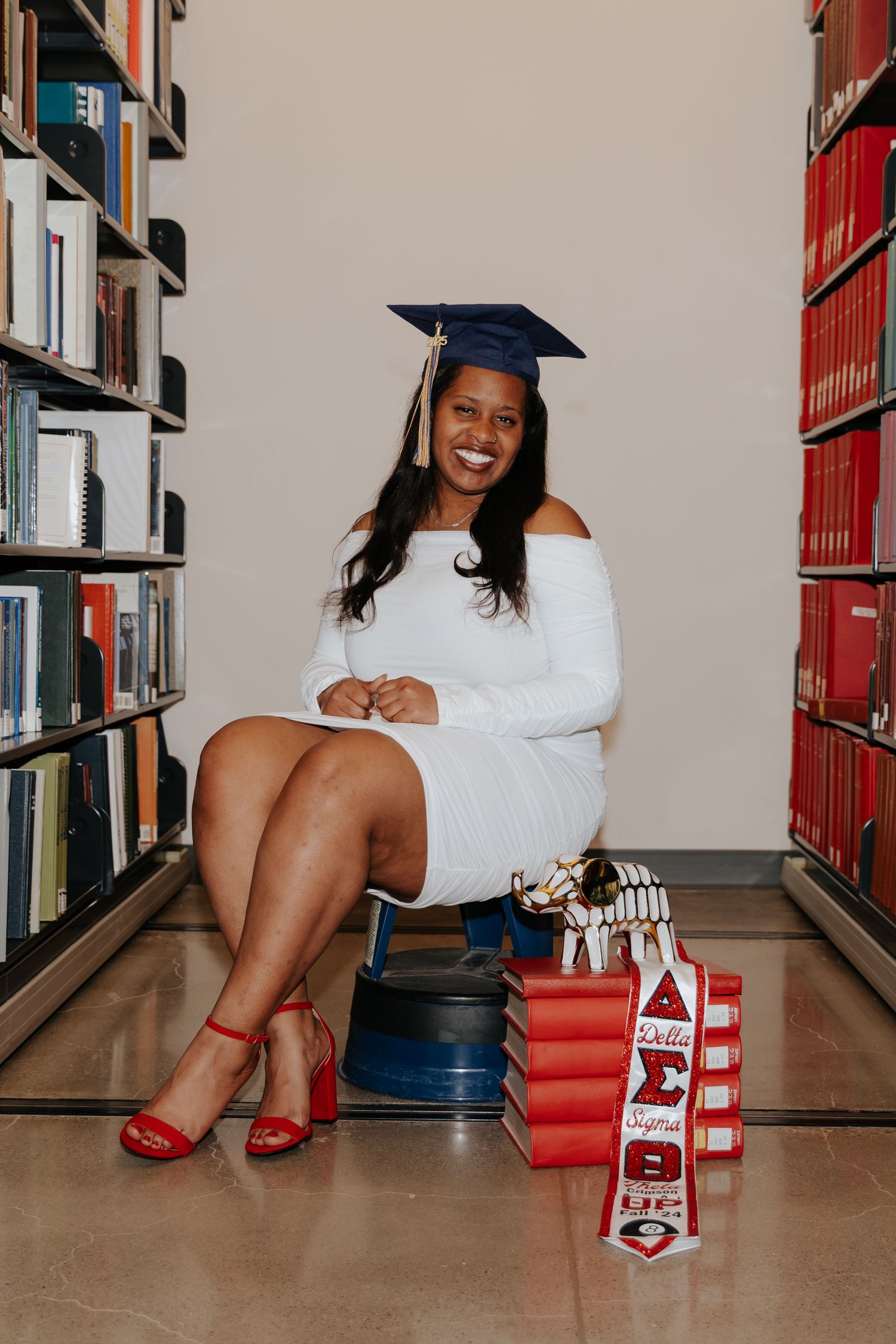 Graduate wearing white dress and graduation cap poses in library with red shoes and decorative letter blocks.