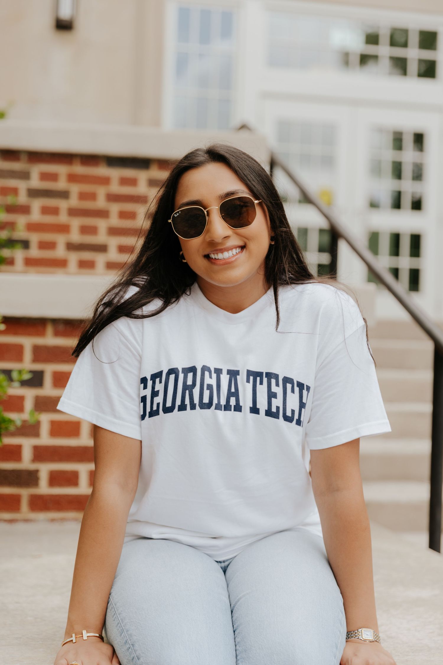 Student wearing Georgia Tech university t-shirt and round sunglasses poses with bright smile in front of brick campus building.
