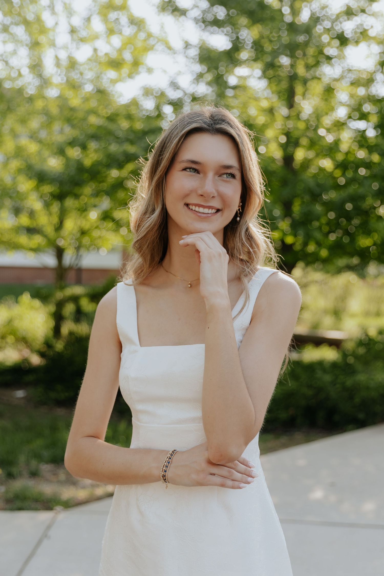 A person in a white sleeveless dress poses outdoors with hand under chin and arm crossed on sunny summer day.