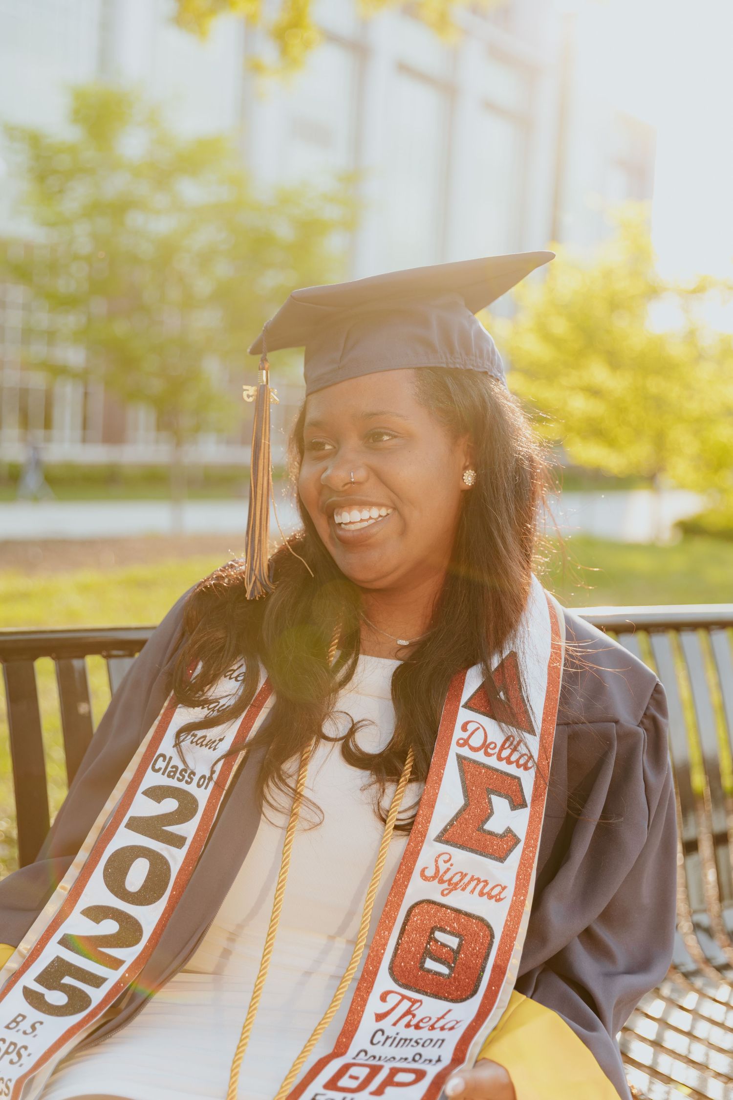 Graduate wearing cap and gown with decorated graduation stoles smiles brightly during outdoor photo session.