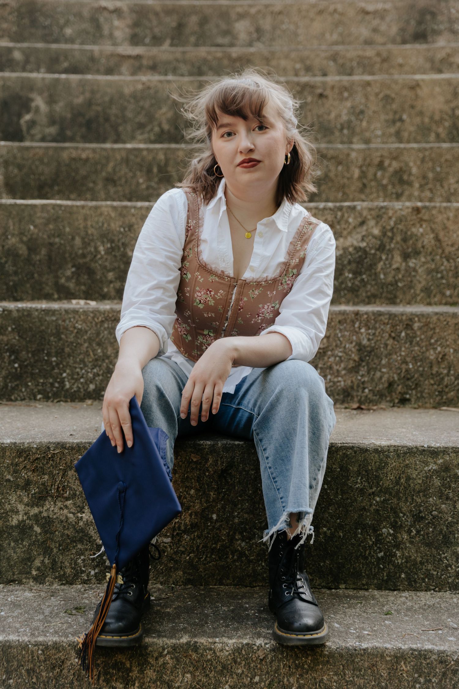 Someone in vintage-inspired outfit with brown corset top, white shirt and jeans sits casually on concrete steps.