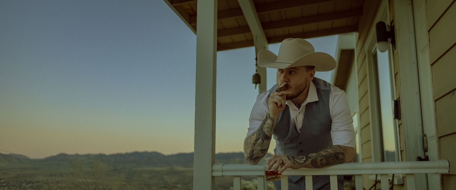 Groom in a cowboy hat and vest leans on a wooden porch railing at sunset with mountains in the background to get that Yellowstone aesthetic