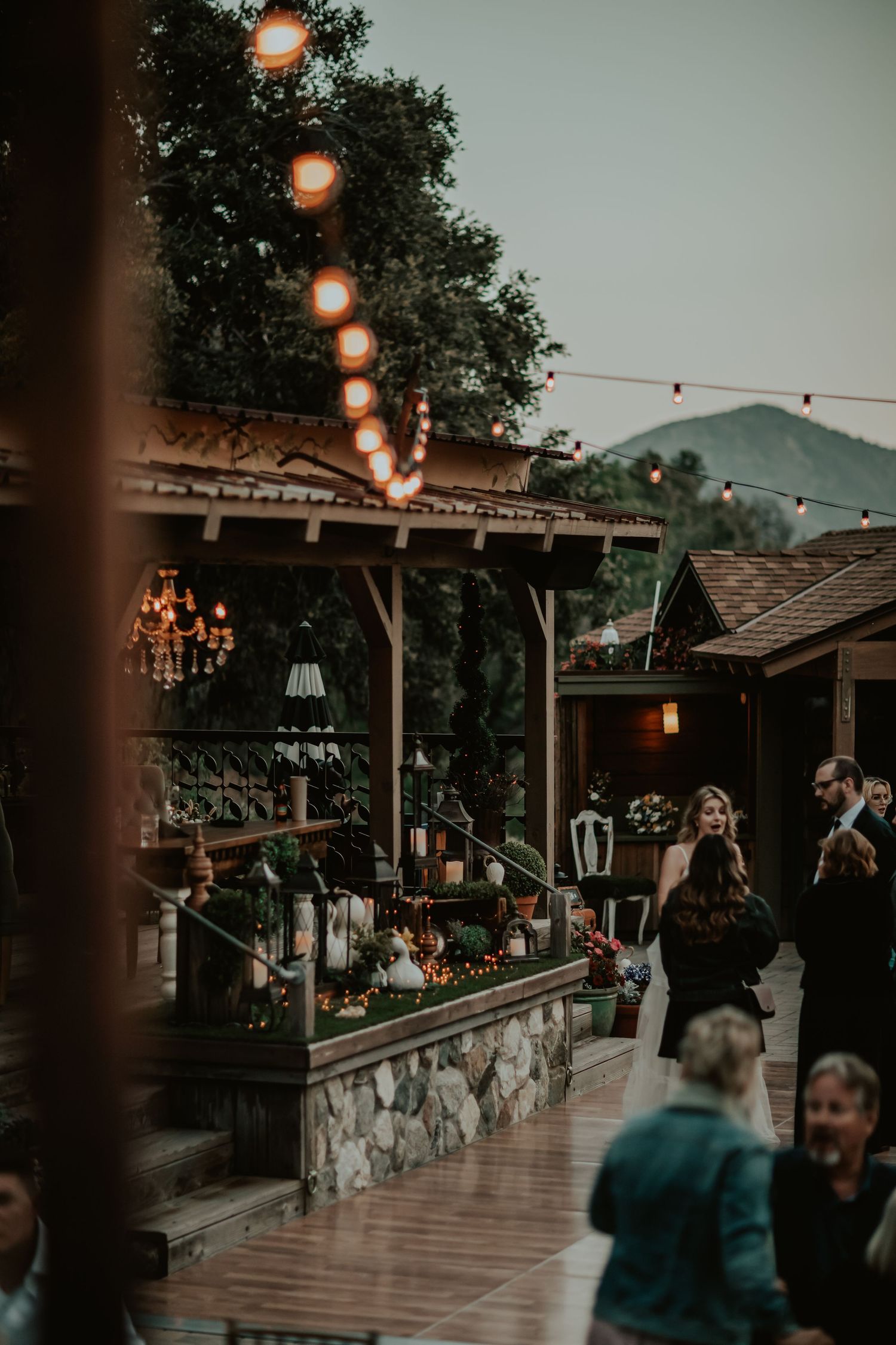 String lights and vintage chandeliers illuminate an outdoor patio deck with stone accents at dusk during a gathering event.