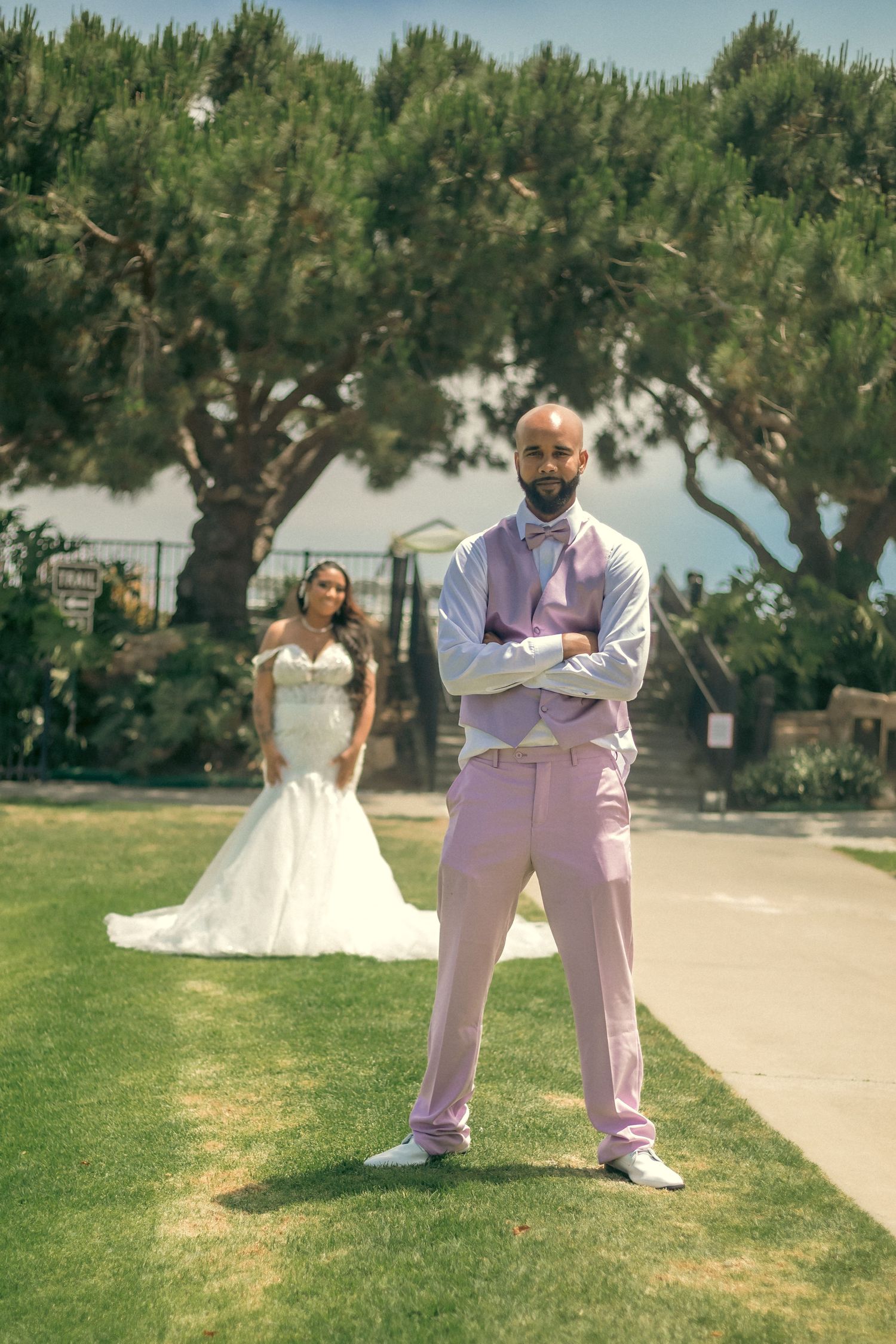A wedding scene on a sunny day with soft pink attire and a white gown against lush green trees and grass.