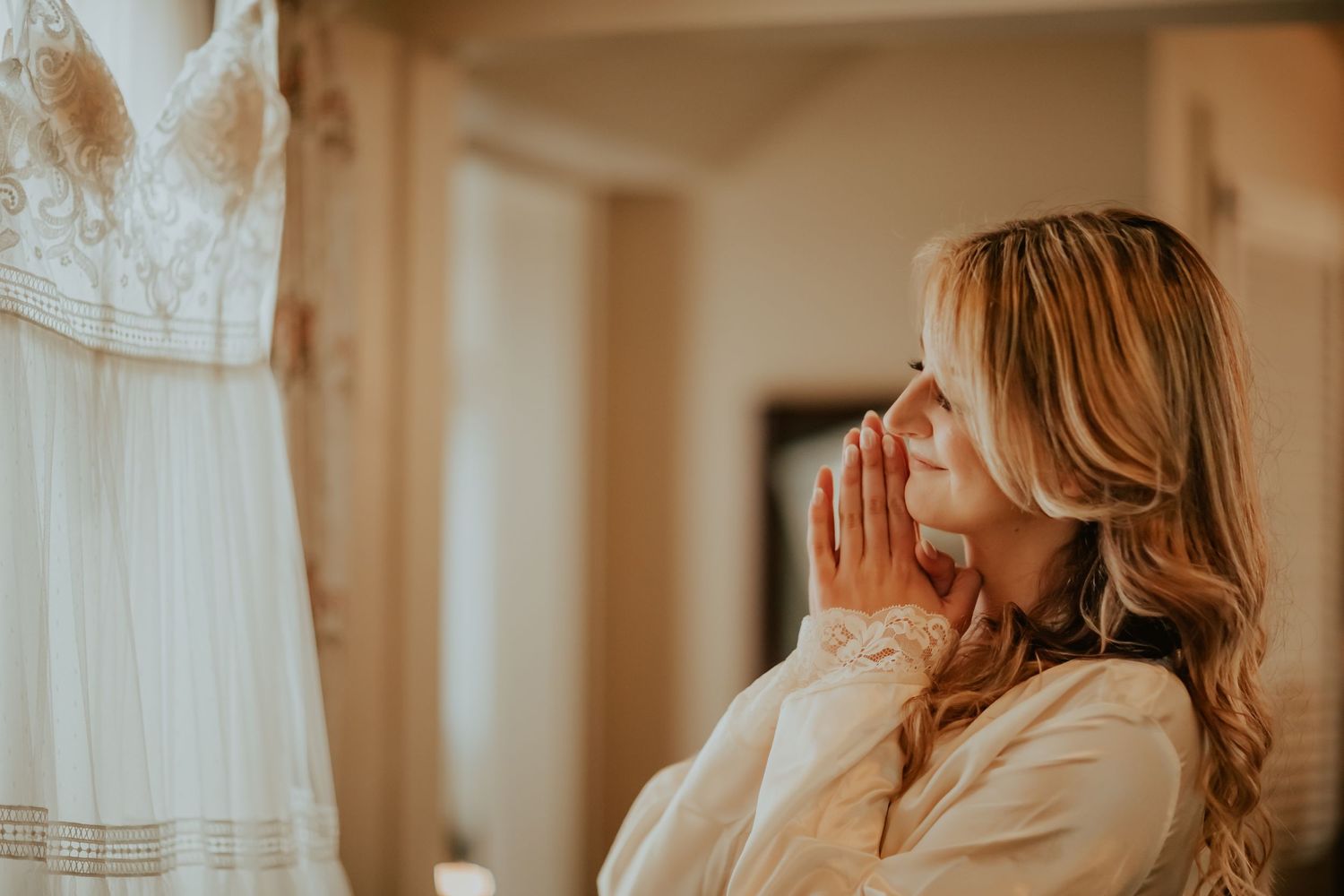 Emotional wedding moment with soft lighting as the bride gazes at white dress hanging nearby.