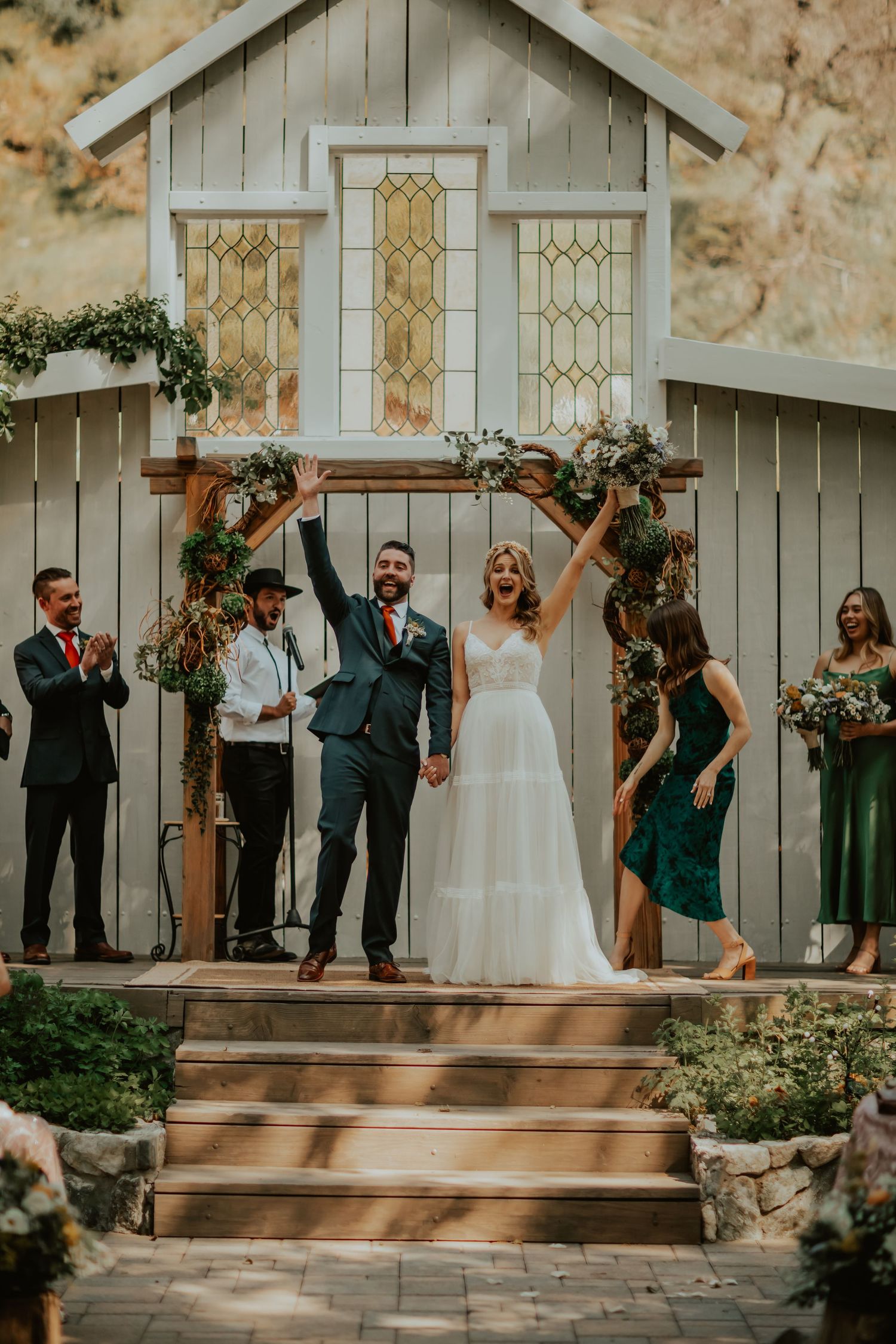 Joyful wedding celebration at rustic outdoor white chapel with wooden stairs decorated in greenery garlands and stained glass windows.