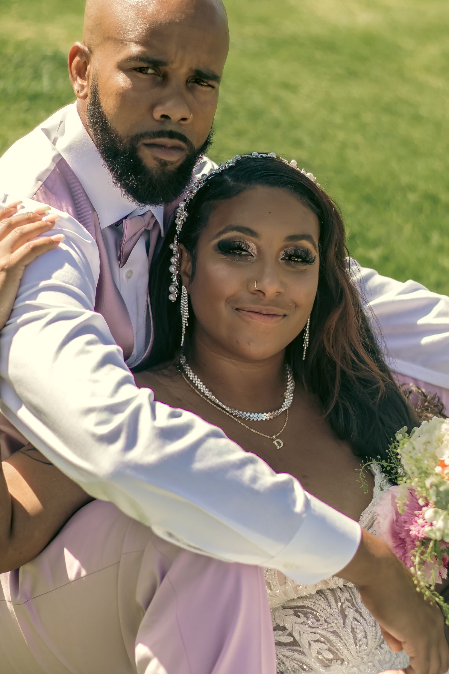 A couple in wedding attire embraces outdoors, with the bride wearing a white lace dress and delicate jewelry sitting a vogue like pose