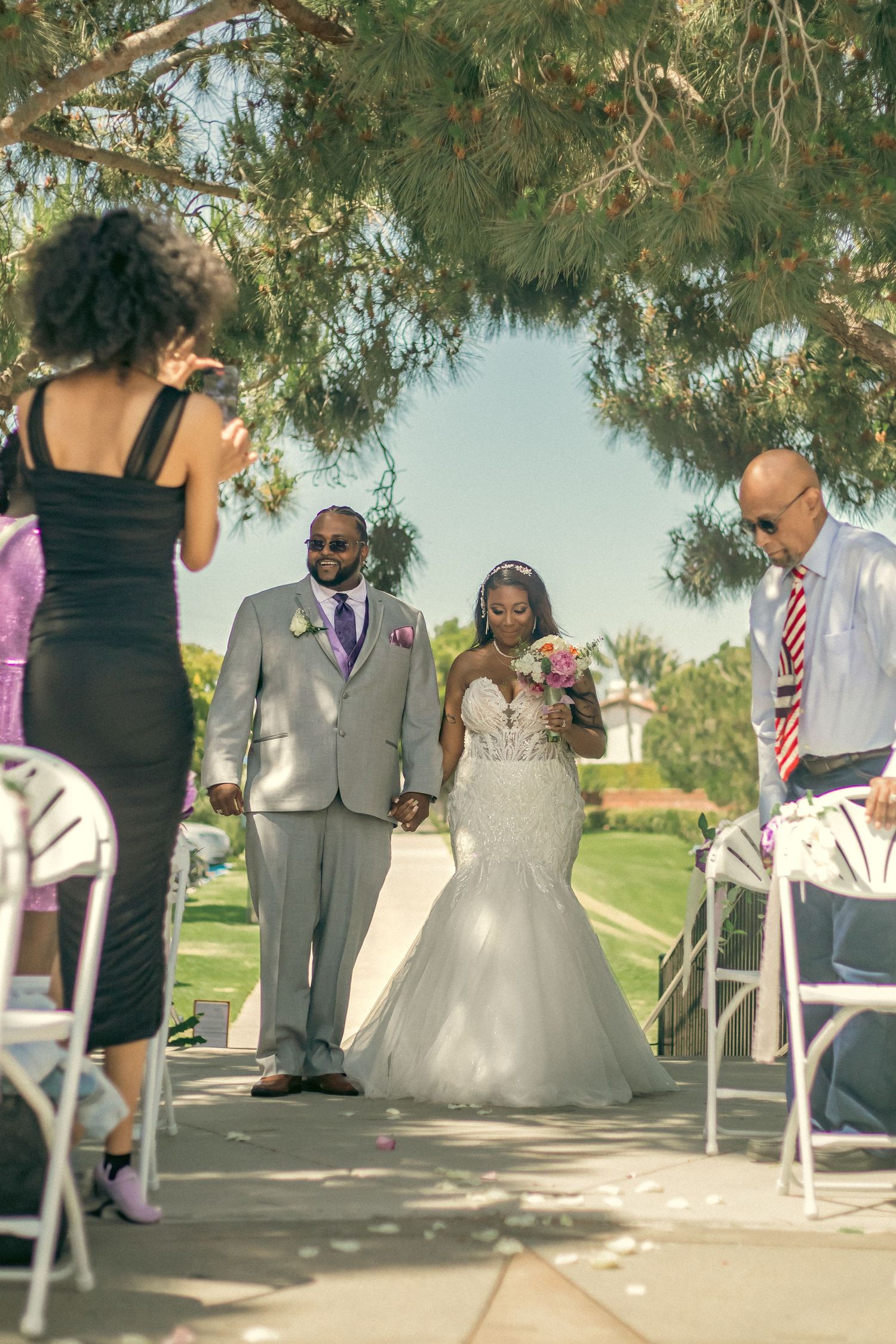 An outdoor wedding ceremony takes place under pine trees with people gathered on a path lined with white folding chairs.