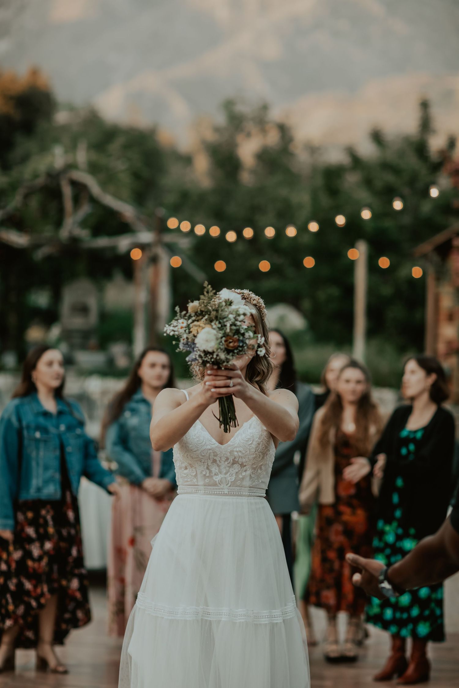 A bride in a white wedding dress holds up her bouquet at dusk with glowing string lights and guests in the background.