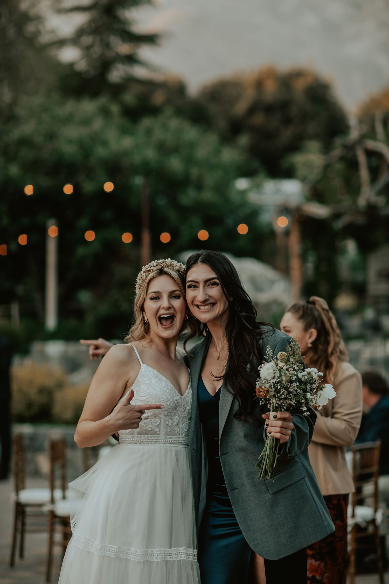 Wedding celebration at dusk with string lights glowing in background and guests holding wildflower bouquets.
