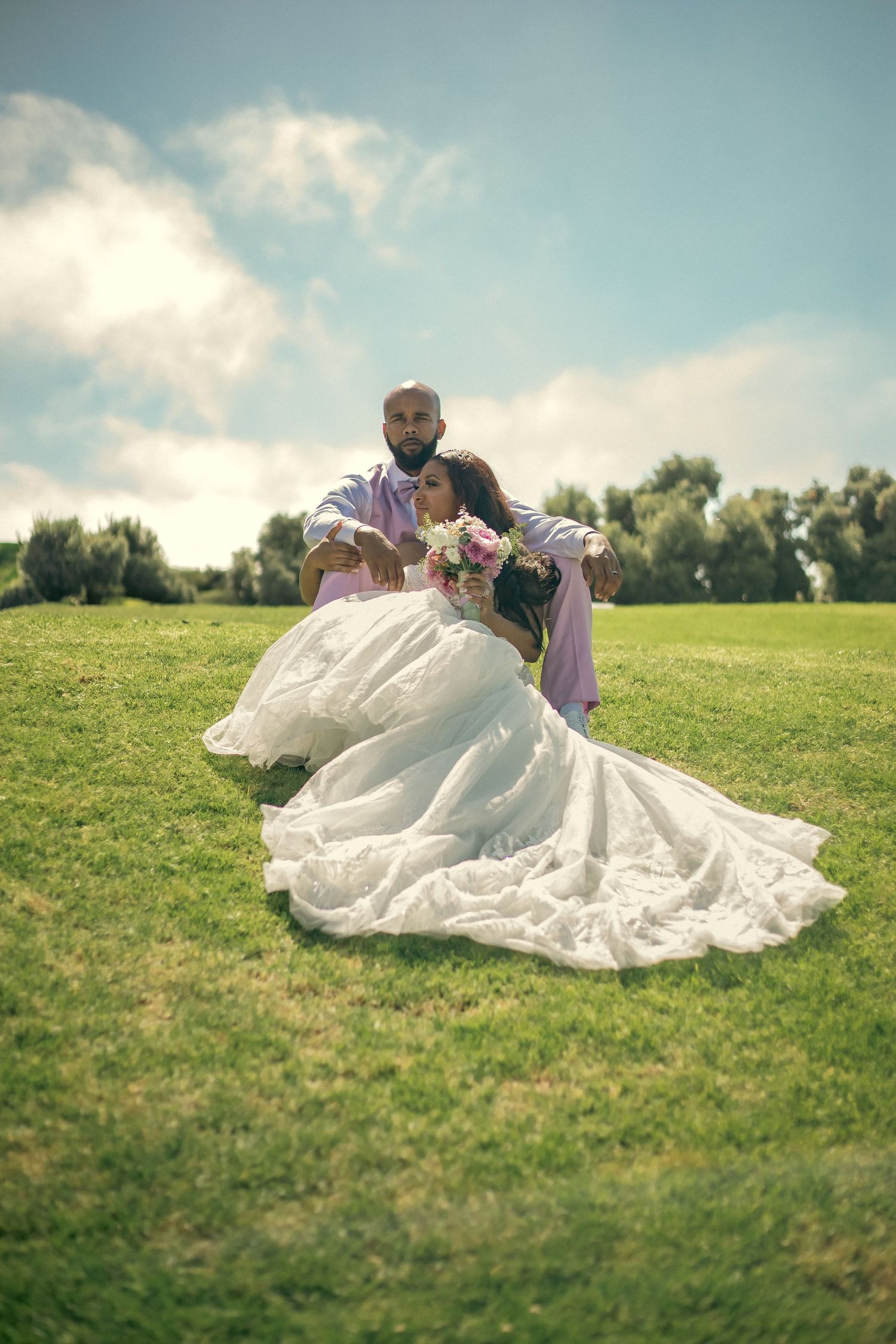 Elegant wedding couple shares romantic moment on green hillside under blue sky and clouds in sunny California