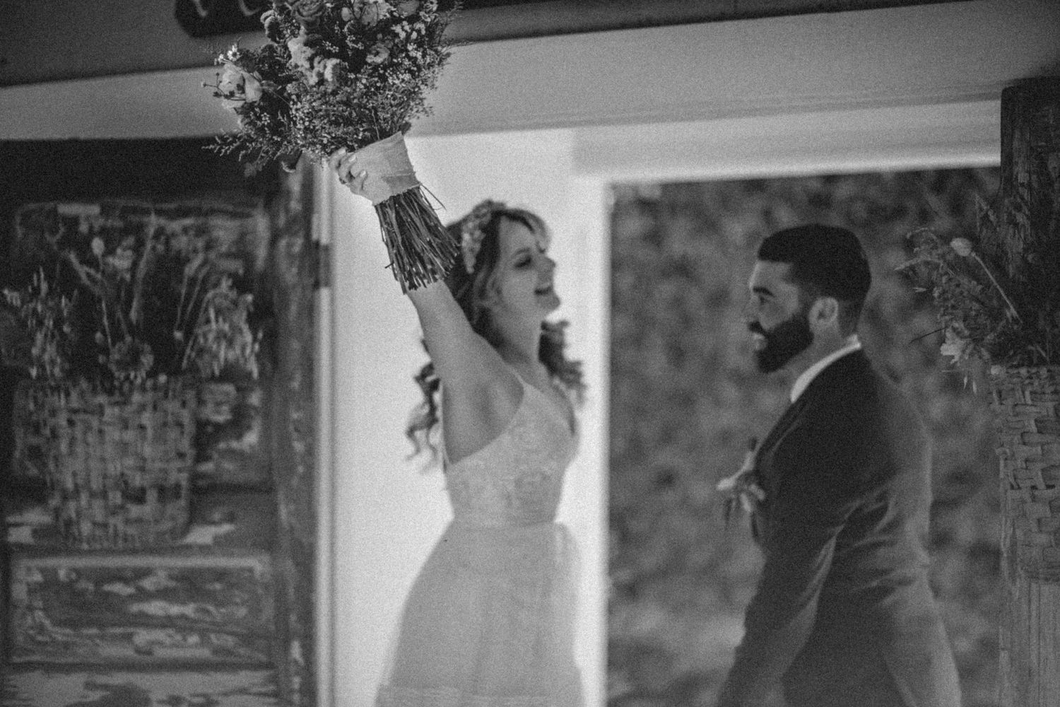 A black and white wedding moment showing an exuberant bride raising her bouquet during a romantic ceremony.