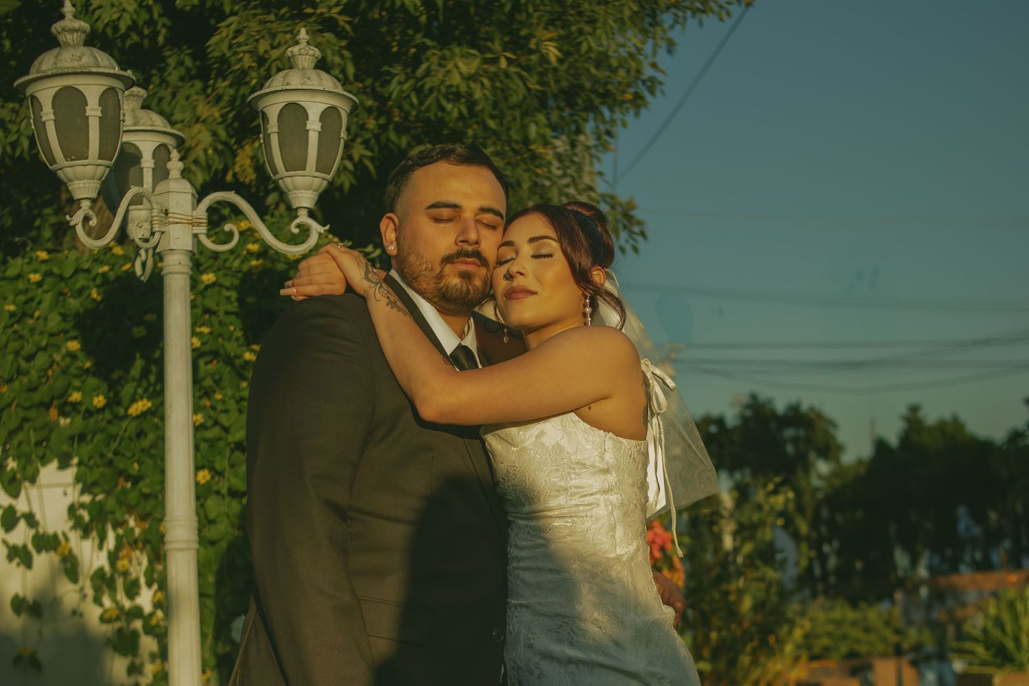 A couple shares a tender moment in their wedding attire at sunset near an ornate lamp post.