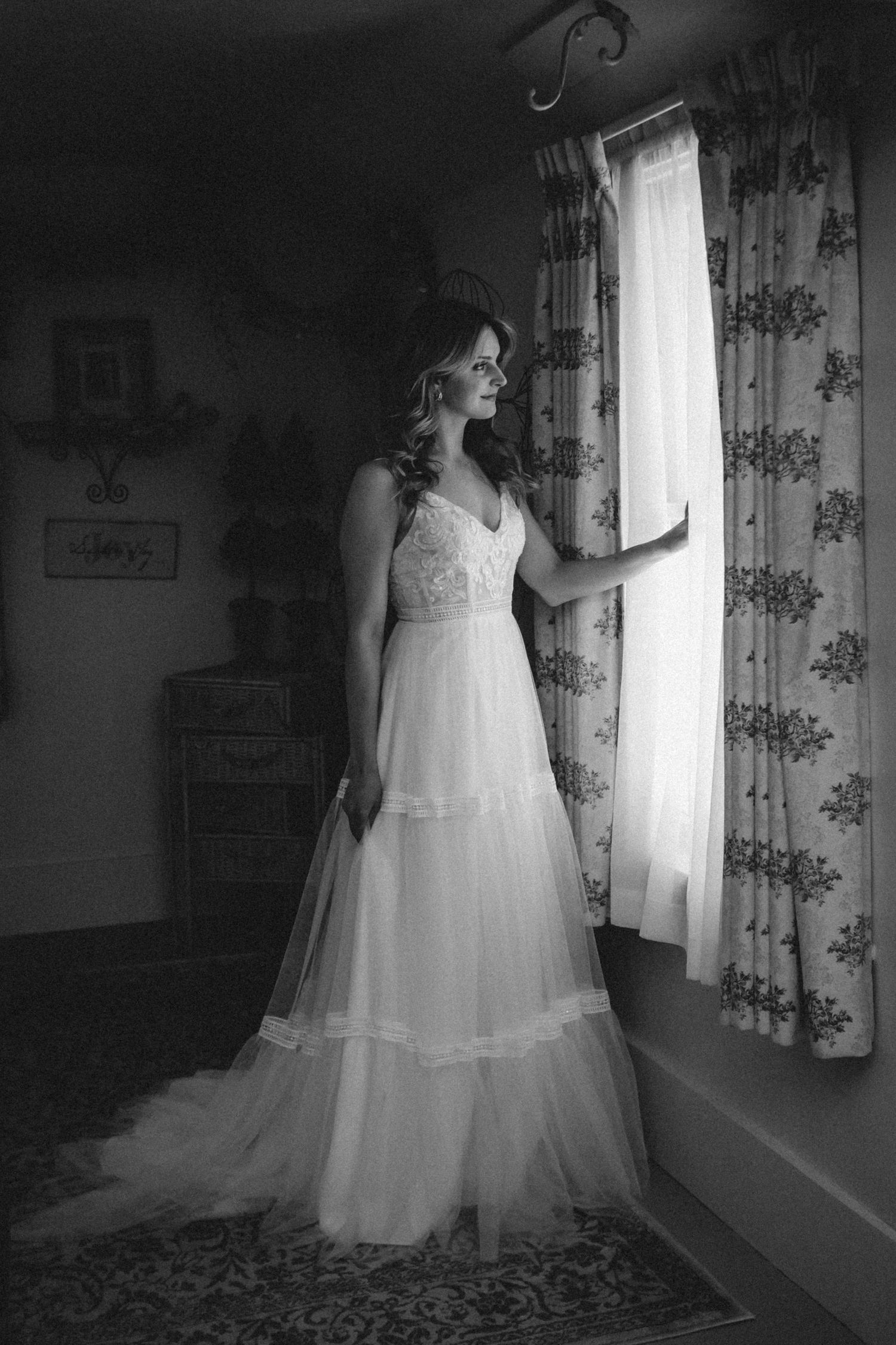 Black and white photo of a bride in a flowing lace wedding gown standing by a window with floral curtains.