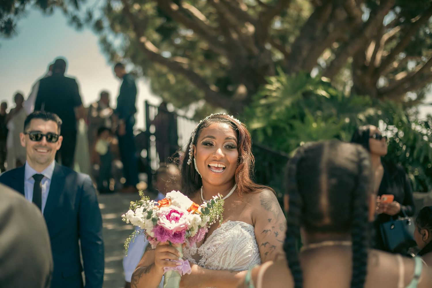 A bride holding a pink and white bouquet smiles joyfully during an outdoor wedding ceremony with palm trees in background.