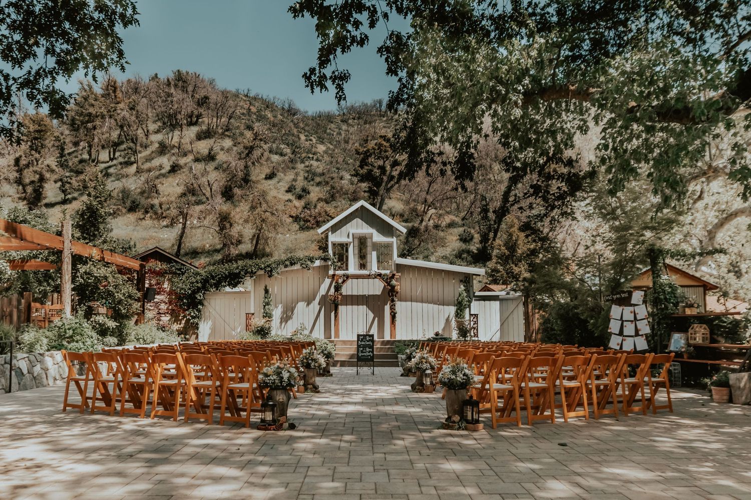 Rustic outdoor wedding venue with wooden chairs arranged in rows facing a white chapel in a mountain setting.