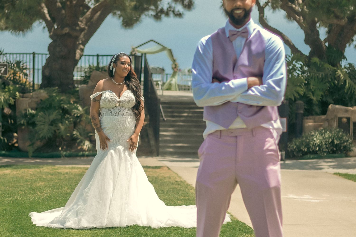 A bride in a lace mermaid wedding dress and groom in a pink suit pose in a sunny garden setting.