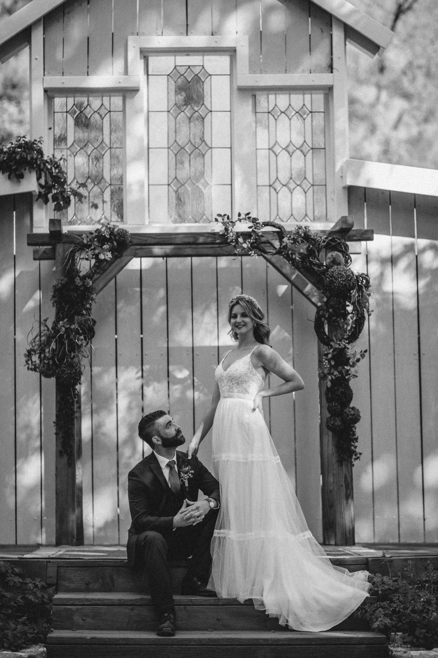 A romantic black and white wedding photo in front of a rustic chapel with stained glass windows and floral arch.
