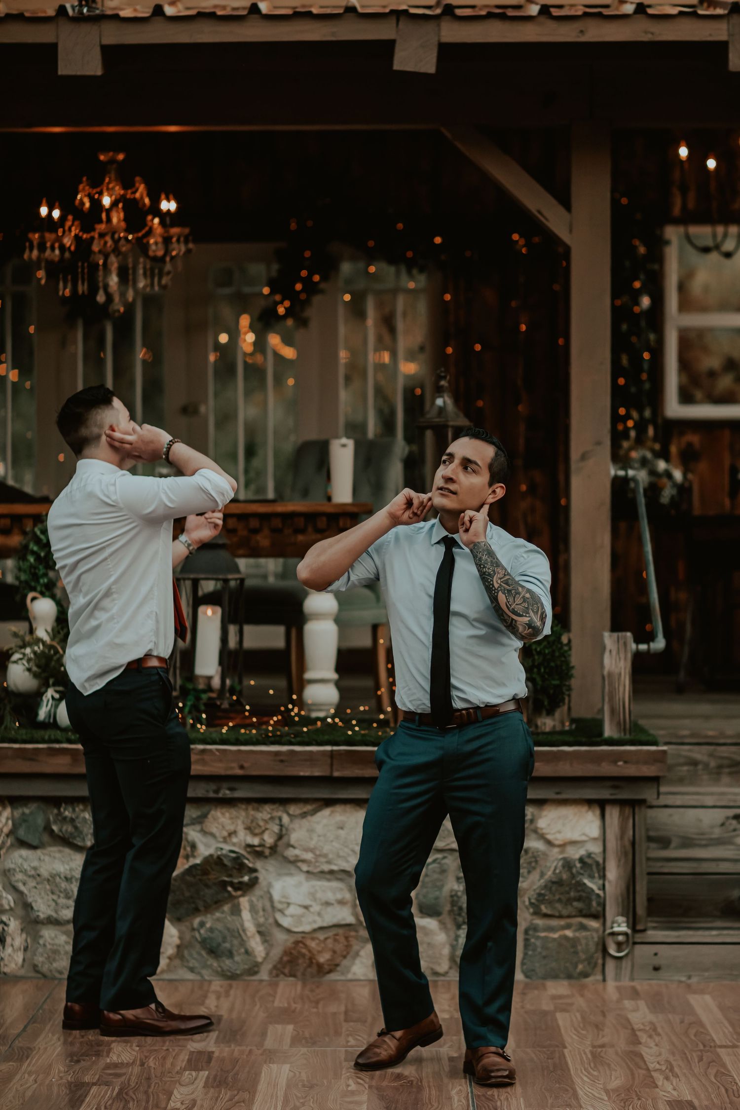 Two guests in dress shirts and ties dance playfully on a wooden floor with a chandelier and stone wall in the background.