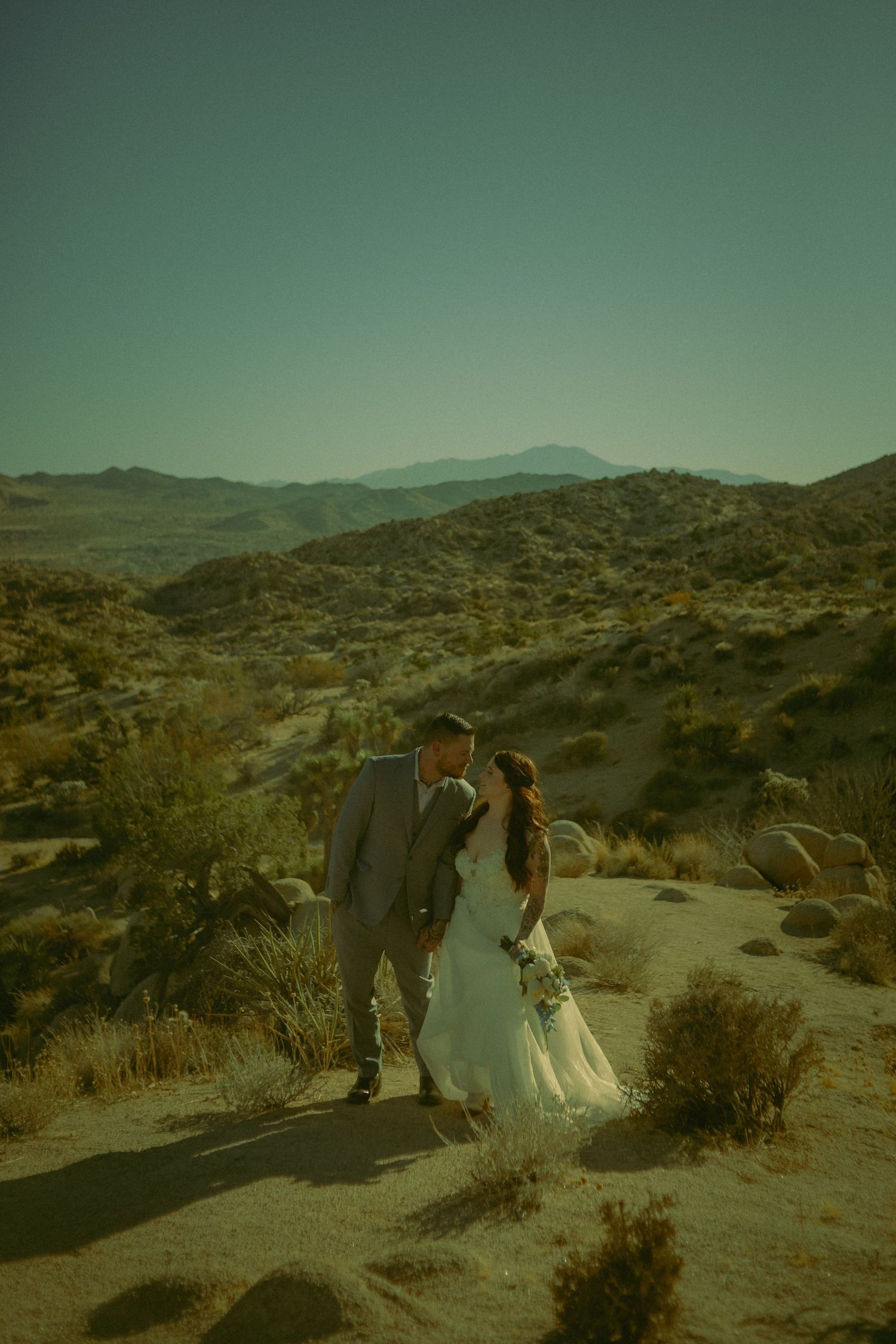 A couple in wedding attire walking together through a desert mountain landscape at sunset.