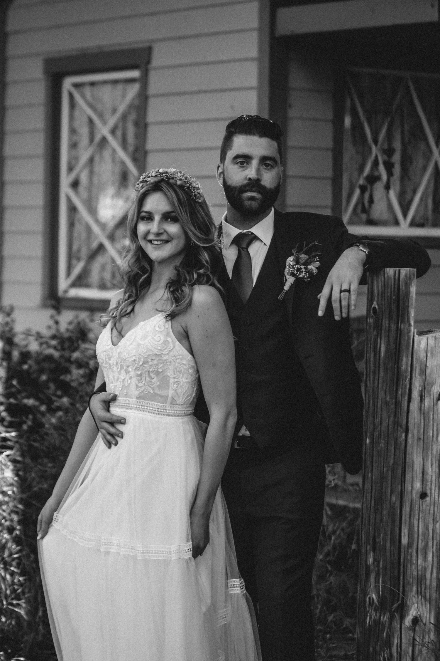 Black and white vogue wedding photo showing bride in lace gown with floral crown and groom in dark suit standing on porch.