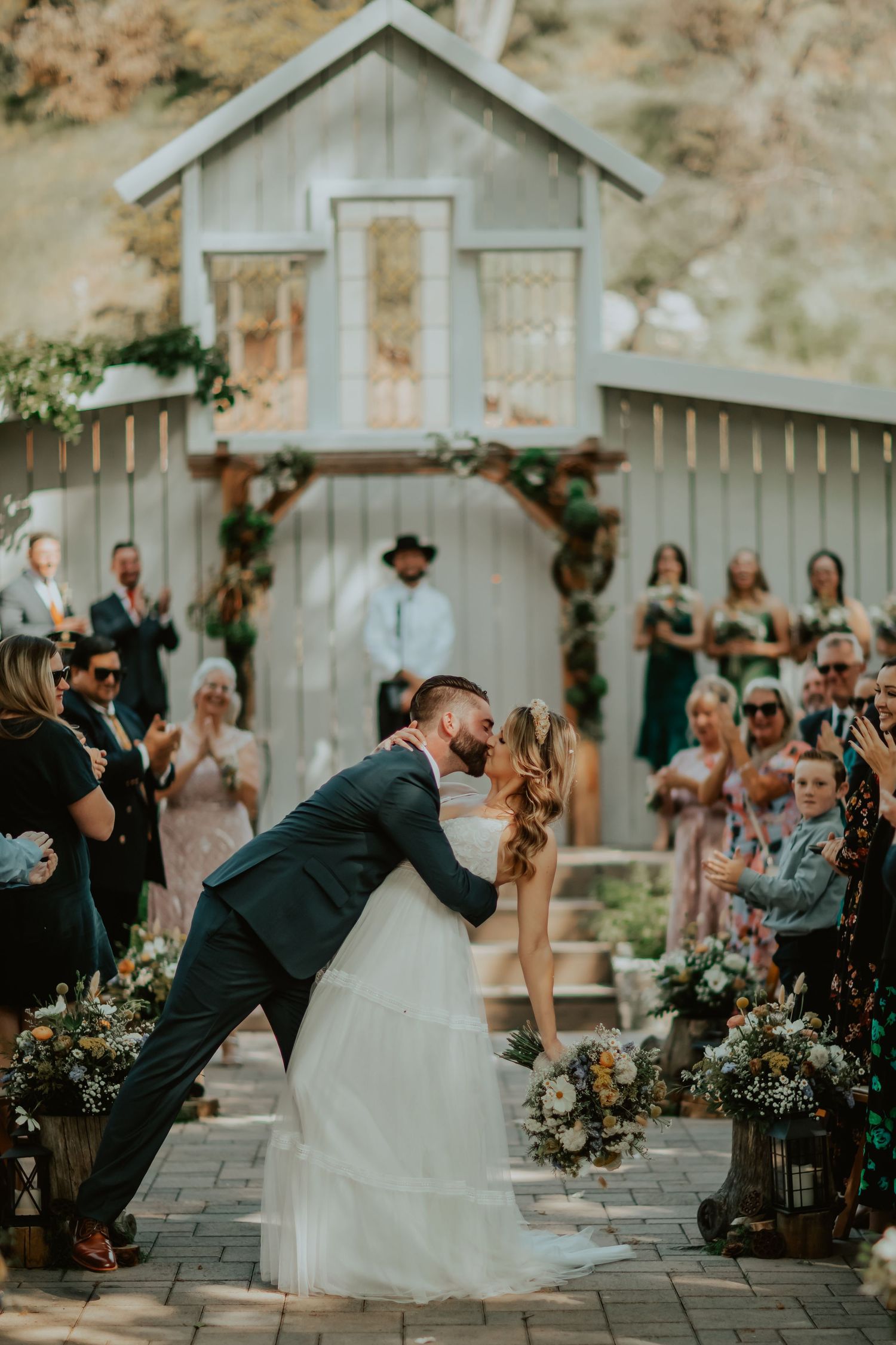 A couple shares a romantic dip kiss at their rustic barn wedding ceremony surrounded by guests and floral decorations.