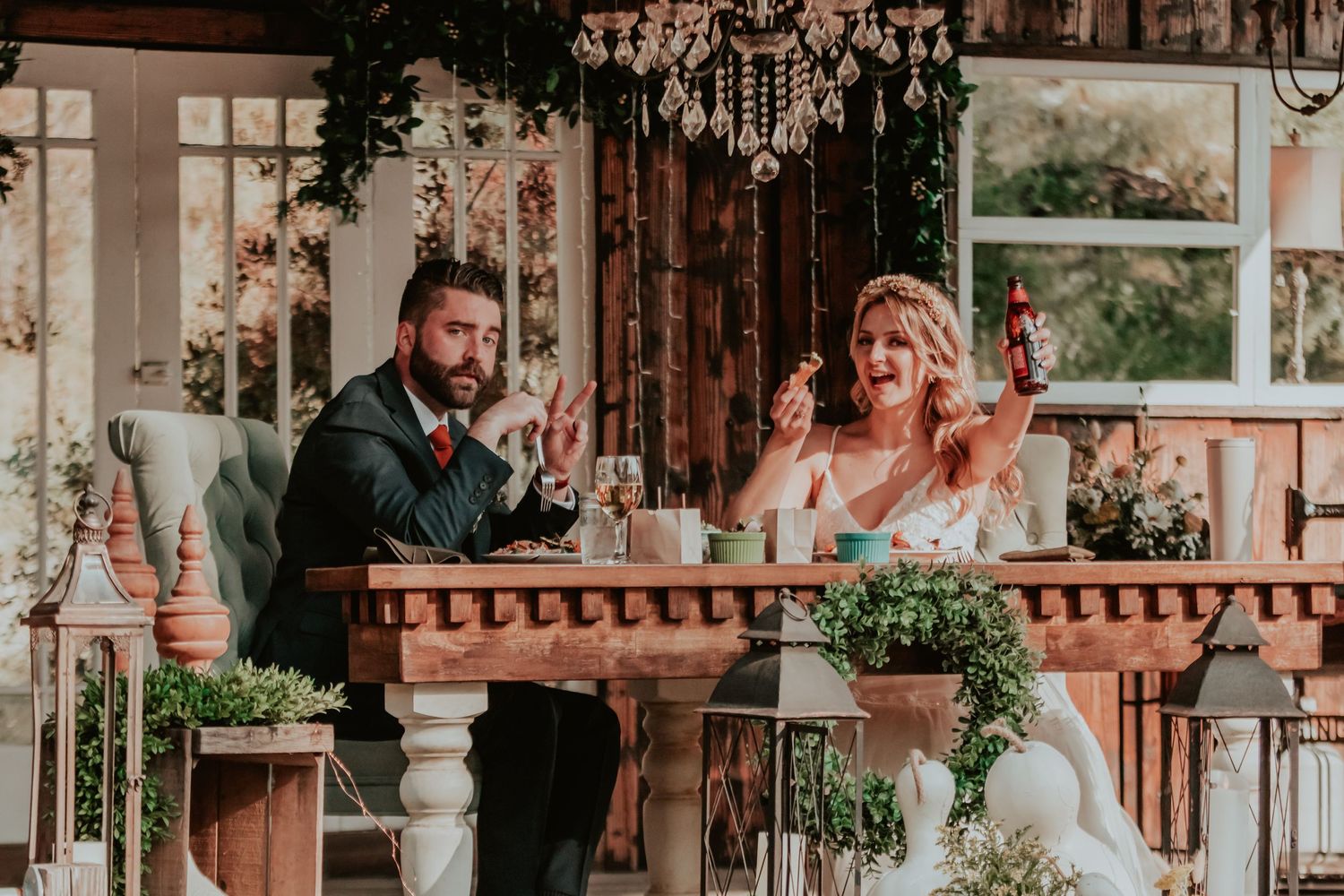 A couple waves excitedly at their wedding reception in a rustic venue decorated with greenery and a crystal chandelier.