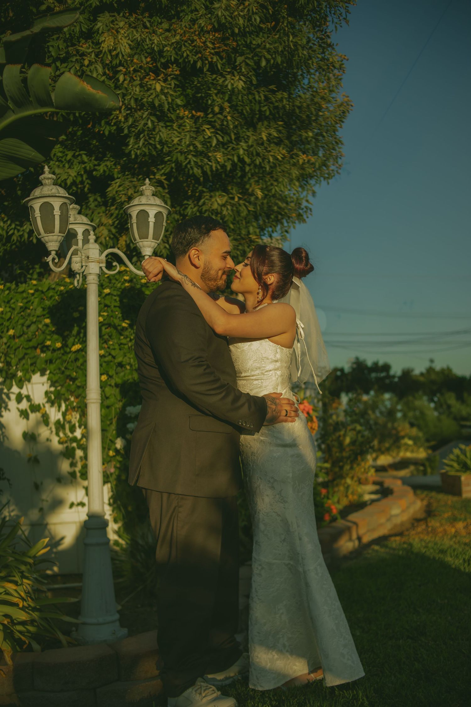A couple embraces in a golden sunset setting with decorative lamp posts and lush greenery in the background.