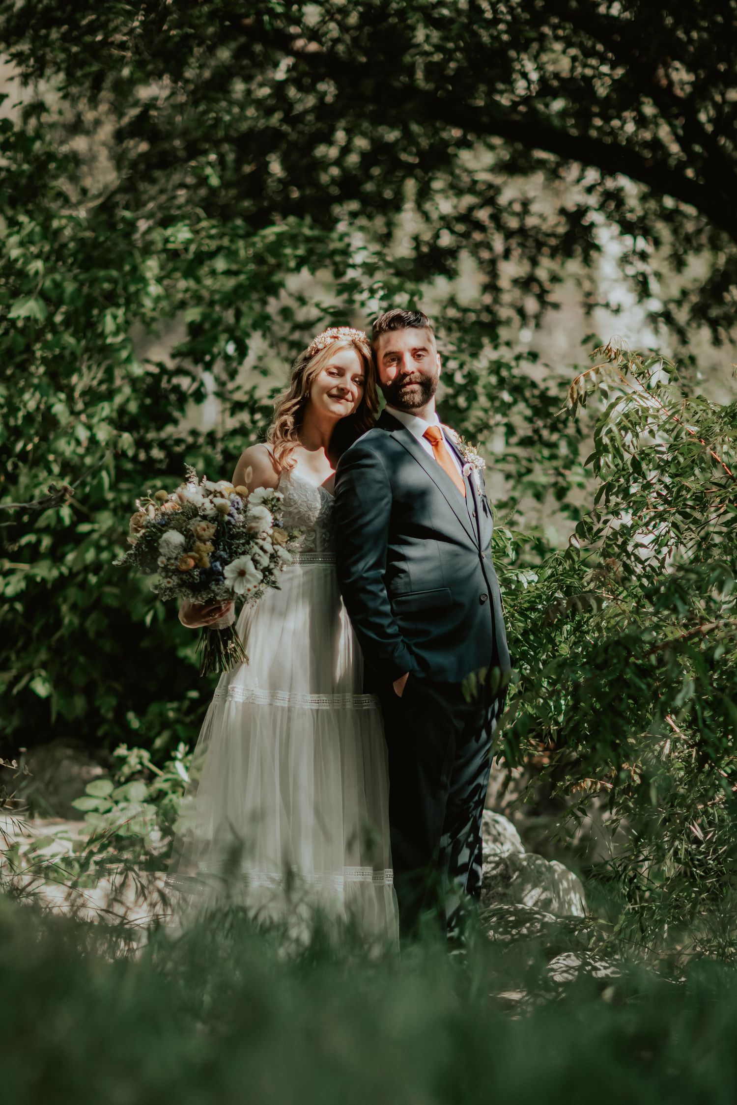 A couple stands together in a lush garden setting, wearing elegant wedding attire surrounded by greenery in California