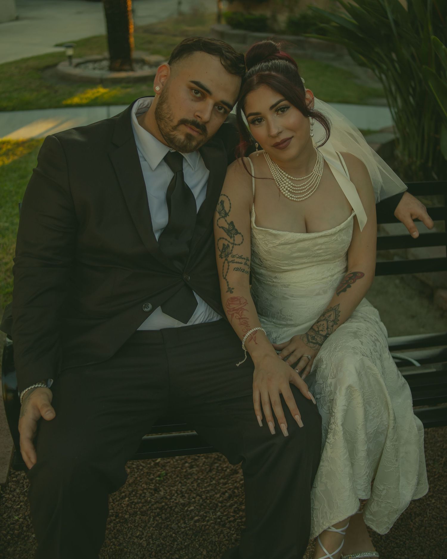 A stylishly dressed bride and groom sit together on a bench in an outdoor garden setting during their wedding.