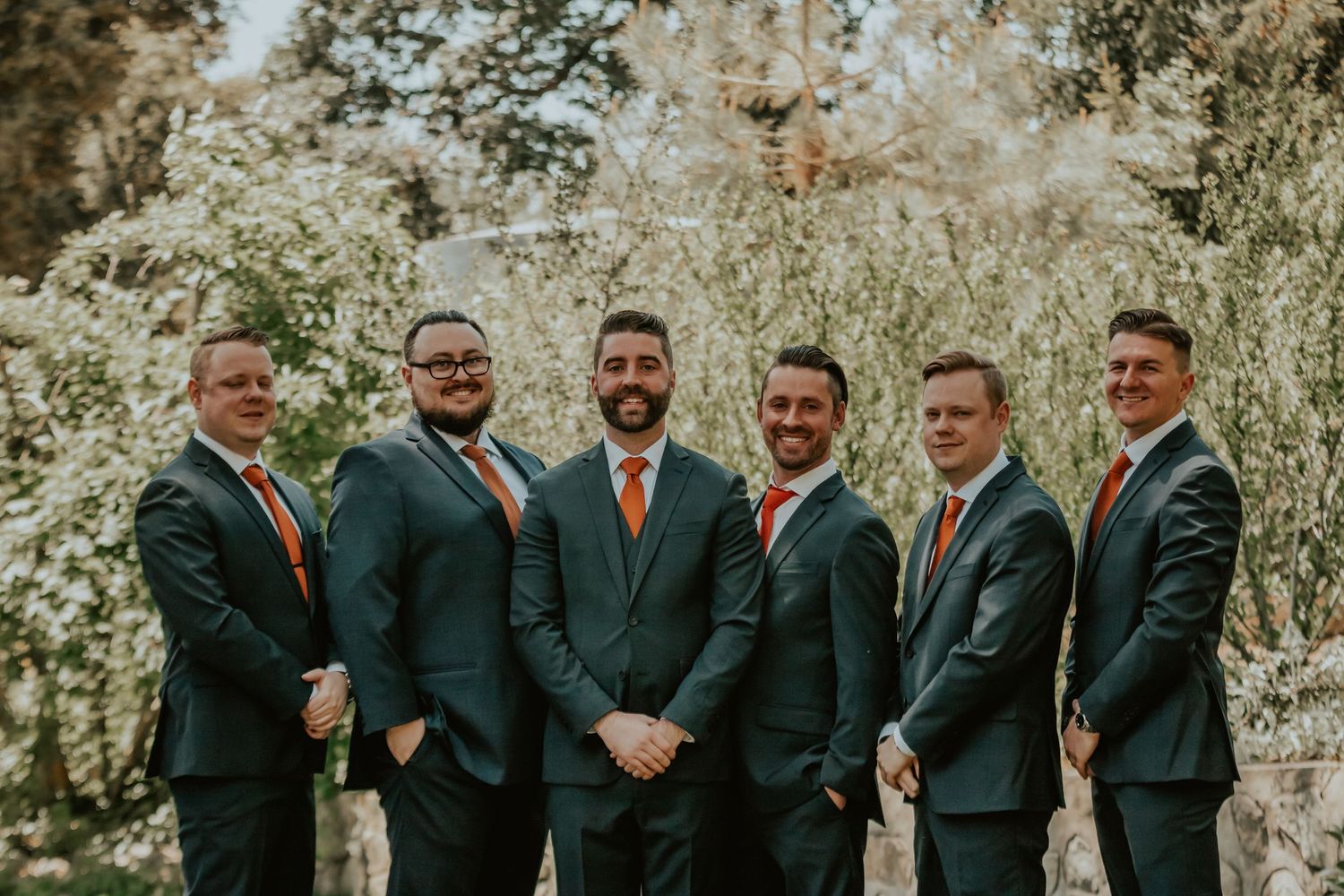 Groomsmen in dark suits with orange ties stand together outdoors in a sunlit wooded setting.