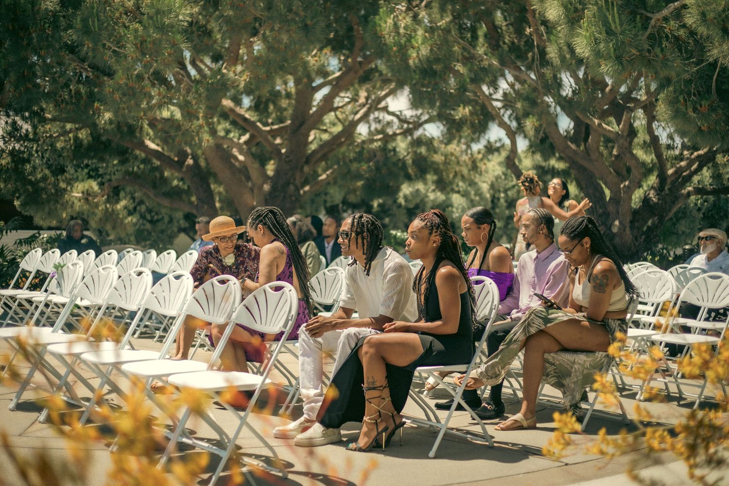 Wedding guests seated on white folding chairs outdoors with floral decorations visible in the foreground.