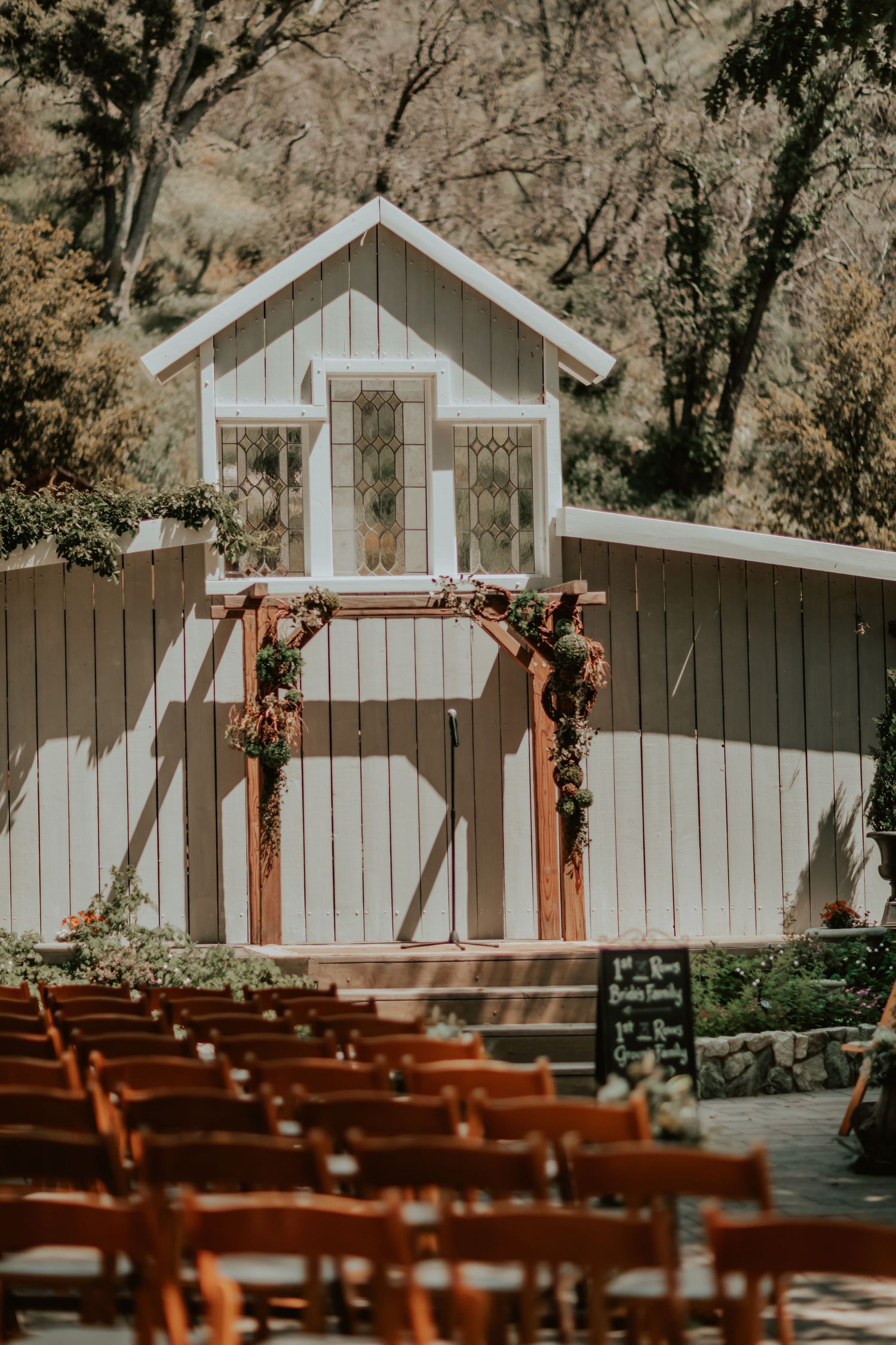 A rustic outdoor wedding venue with a white chapel backdrop and wooden arch decorated with autumn flowers and empty chairs.