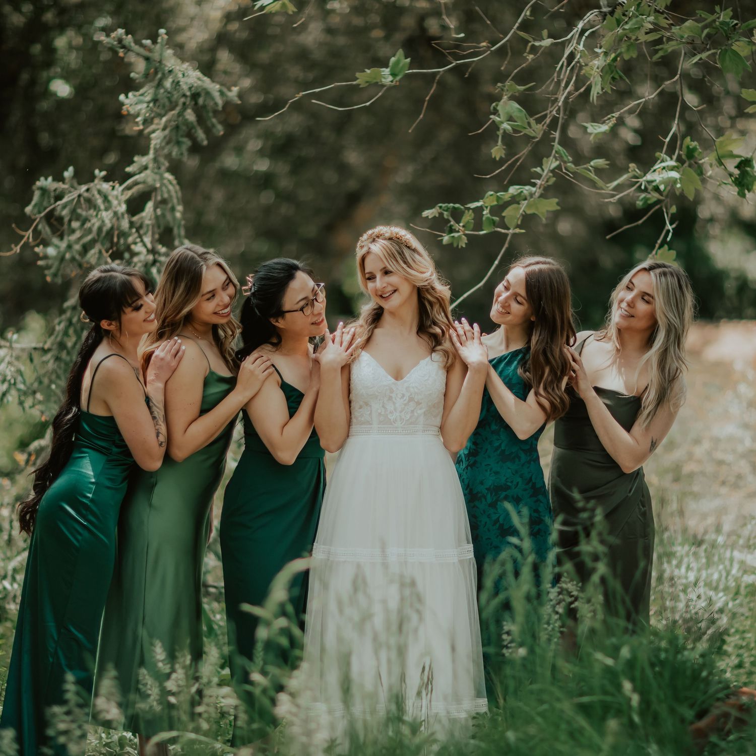 A bridal party in emerald green dresses shares a candid moment in a sunlit garden setting happily surrounding the bride