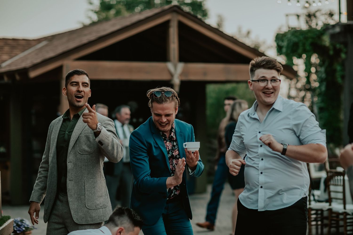 Three friends enjoy beverages together at The Homestead wedding with a wooden structure in the background.