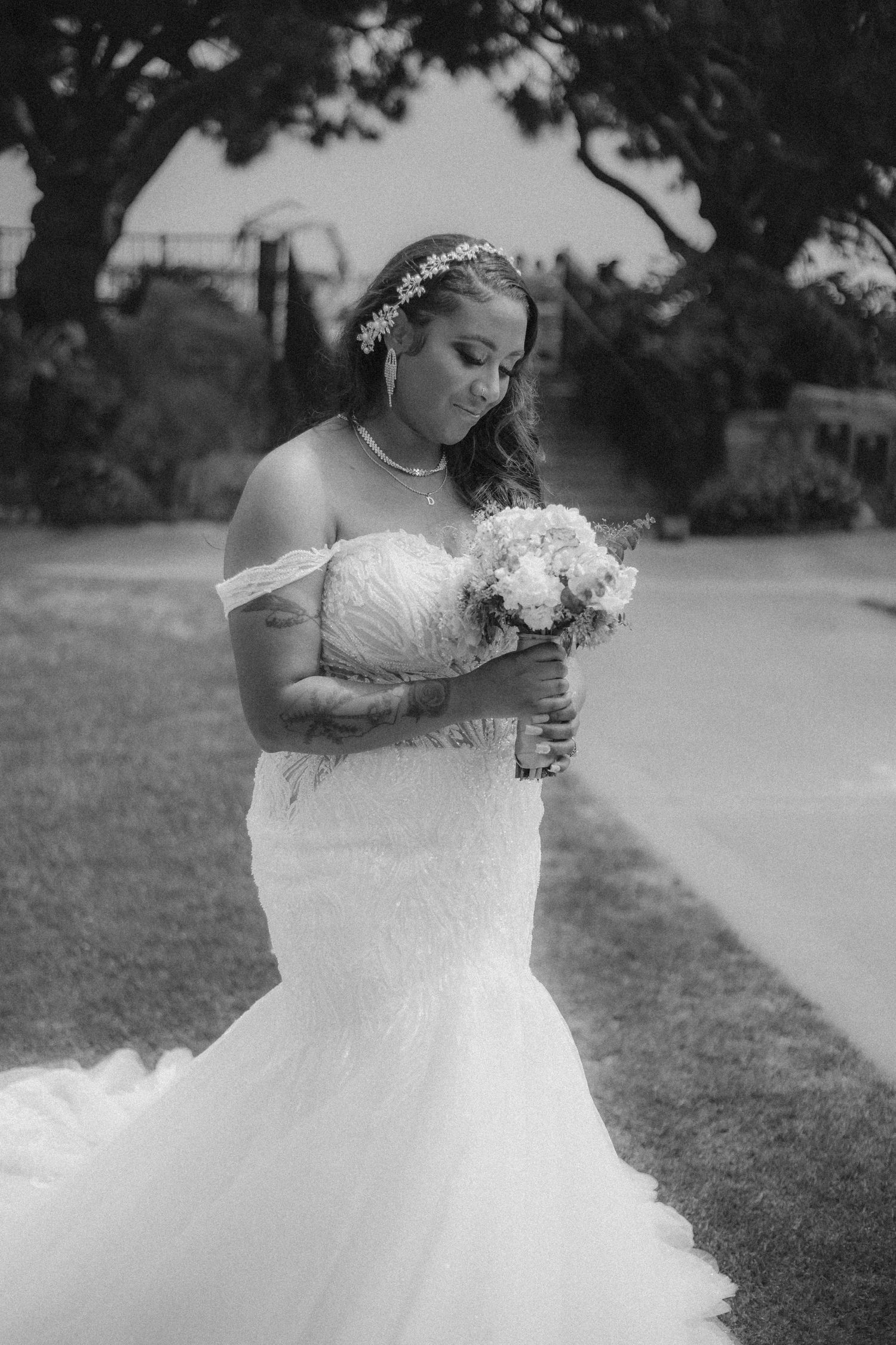 Black and white wedding photo of a bride in an off-shoulder mermaid gown holding a bouquet in a garden setting.