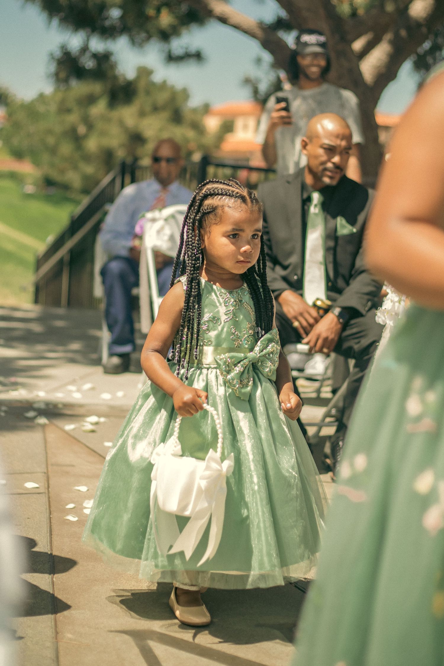 Flower girl with braided hair and white purse walks along outdoor path at wedding venue in California.