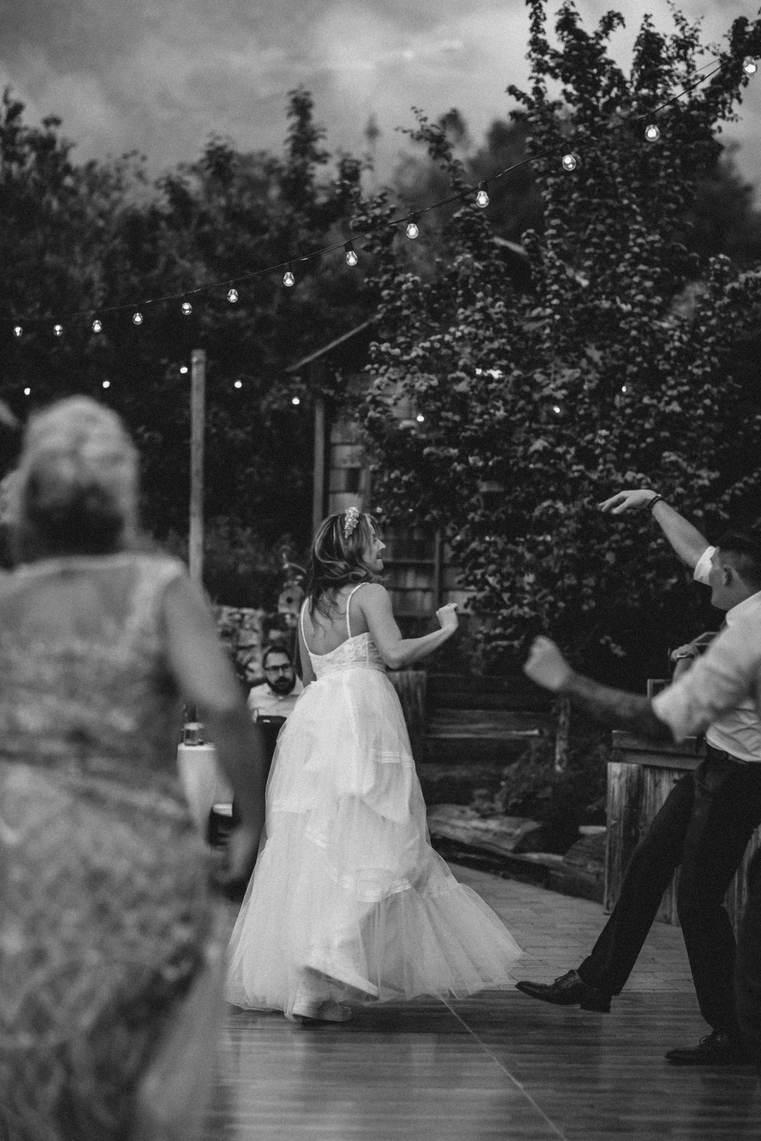 A black and white wedding photo of people dancing on a deck under string lights at an intimate outdoor reception party.