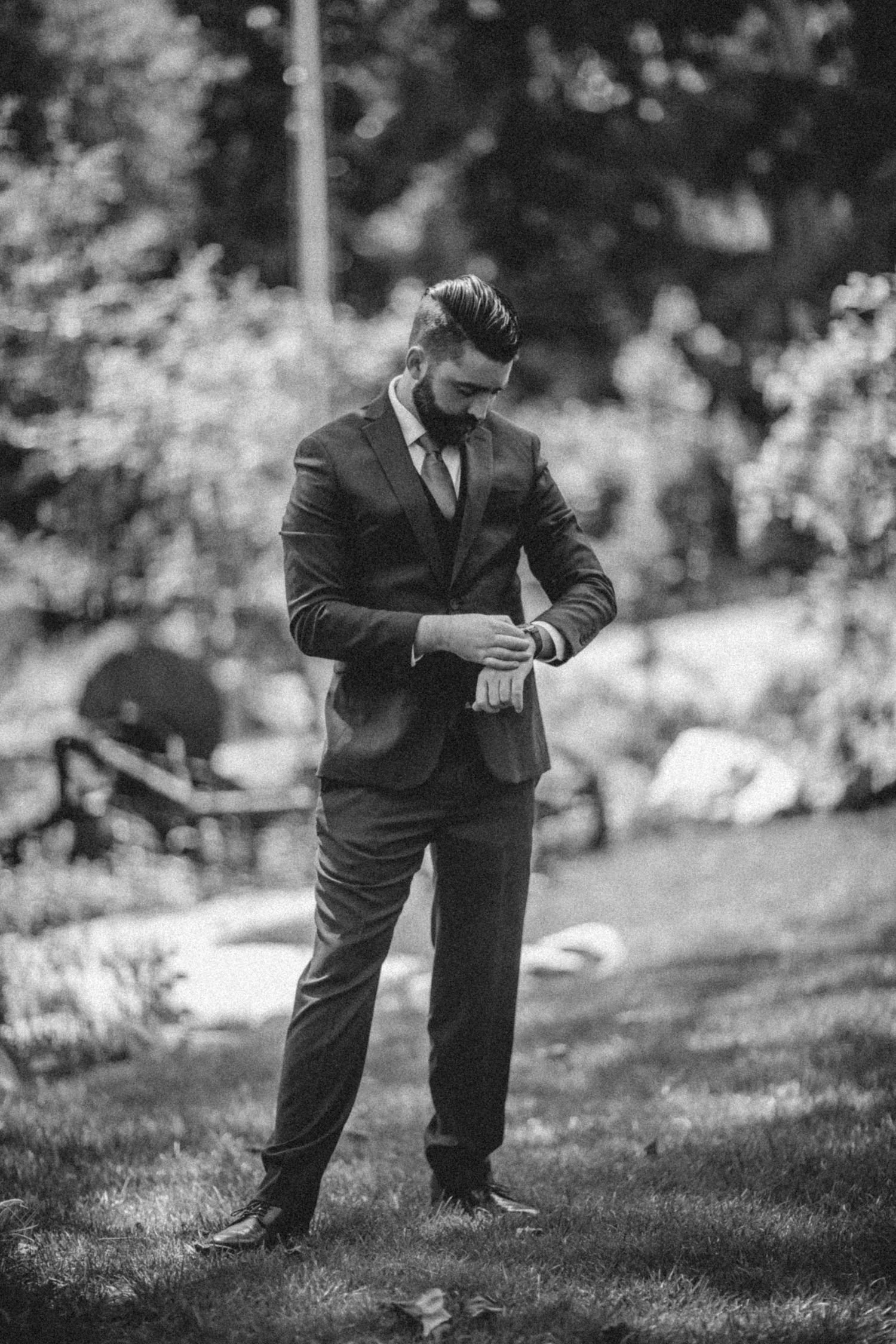 Groom in a dark suit checks his watch while standing on a grassy lawn in black and white photography.