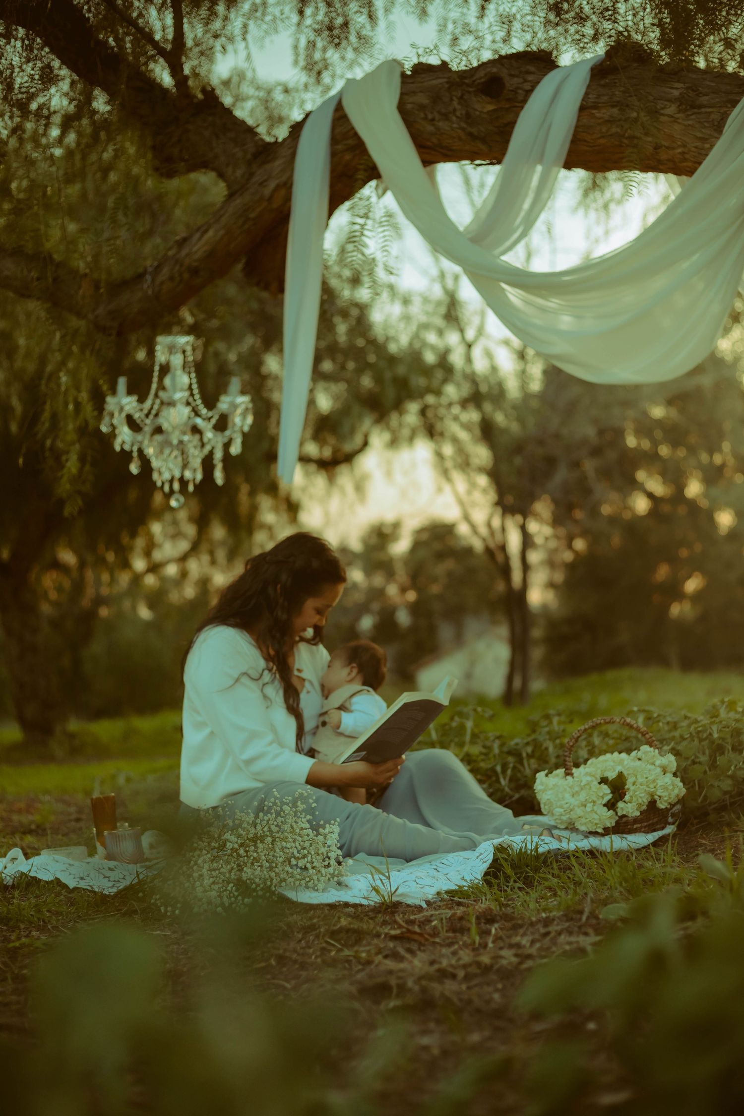 Mother reading under white fabric draping from trees, crystal chandelier, and flowers creating a whimsical atmosphere.