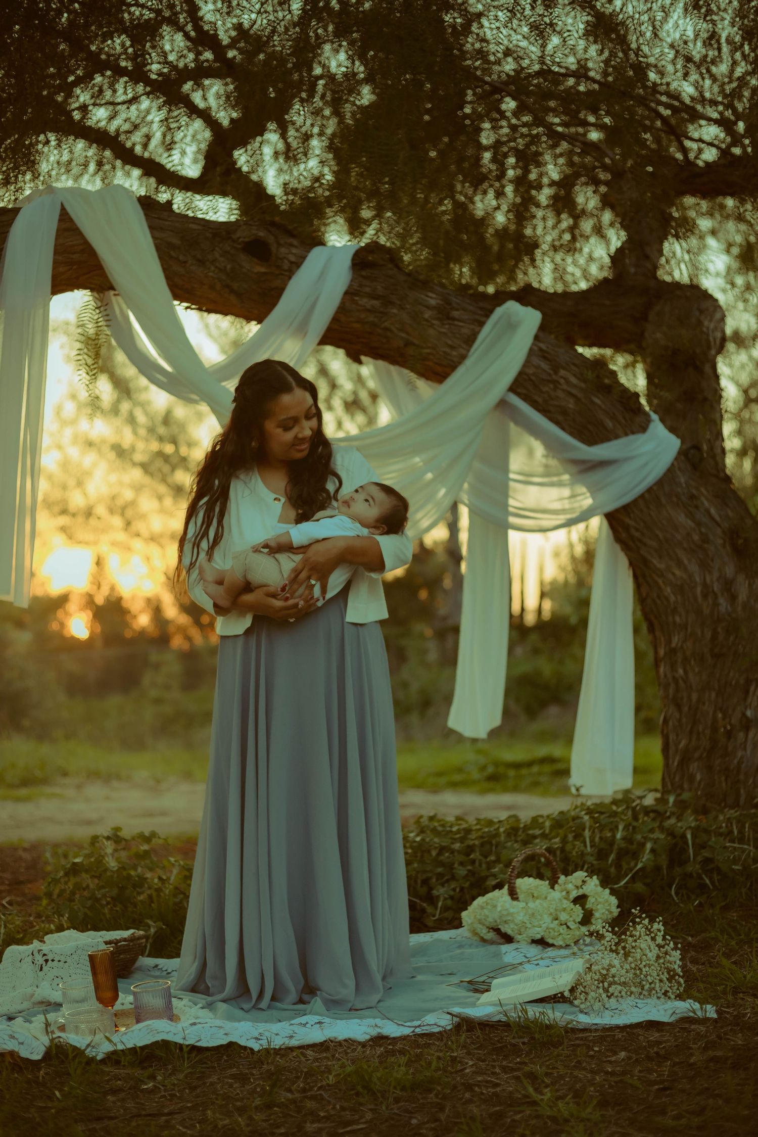 SoCal mother in a flowing blue dress holds an infant beneath white fabric draped from a tree at sunset in a dreamy outdoor mini session.