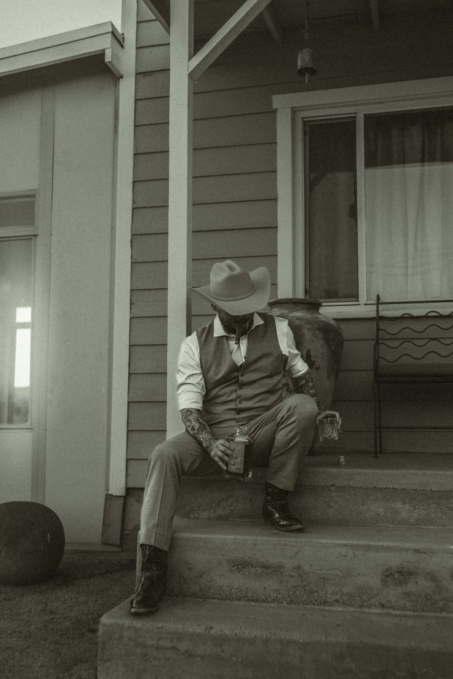 Groom in vintage western attire sits on porch steps of a desert house in a black and white photograph before elopement ceremony.