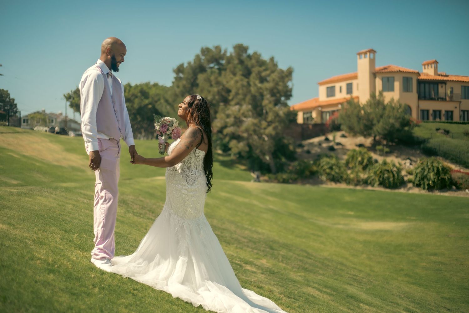 Wedding couple standing on a green hillside lawn with a Mediterranean style villa in the background.