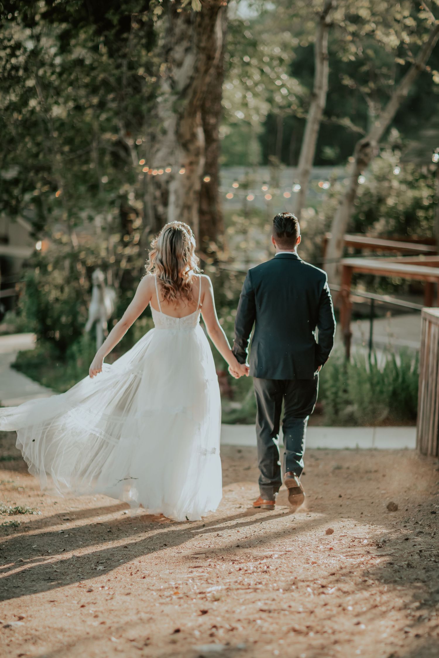 A couple in wedding attire walk hand in hand down a rustic dirt path surrounded by trees and natural scenery.
