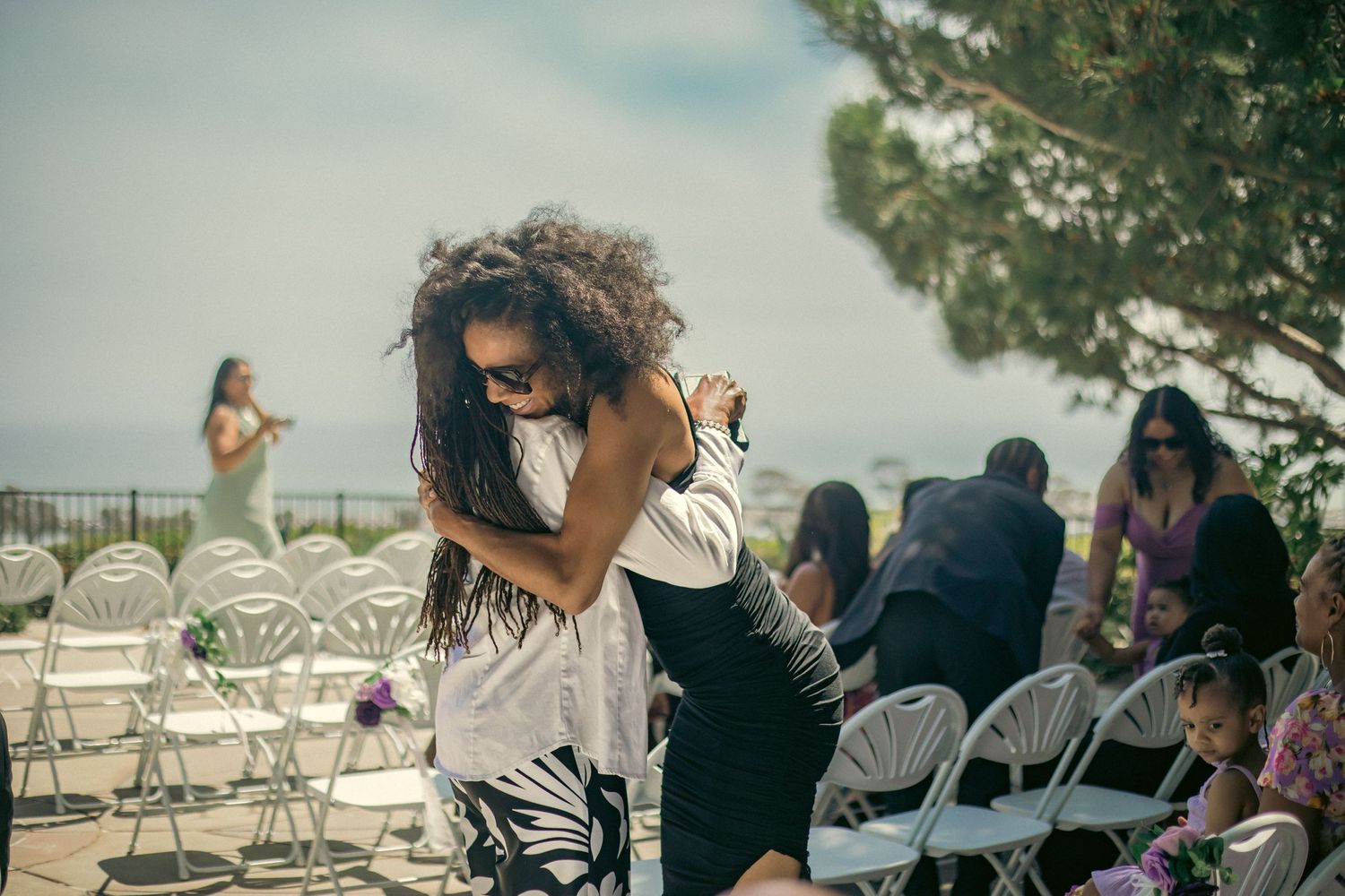Two people embrace in an emotional moment at an outdoor event with white chairs and guests in the background.
