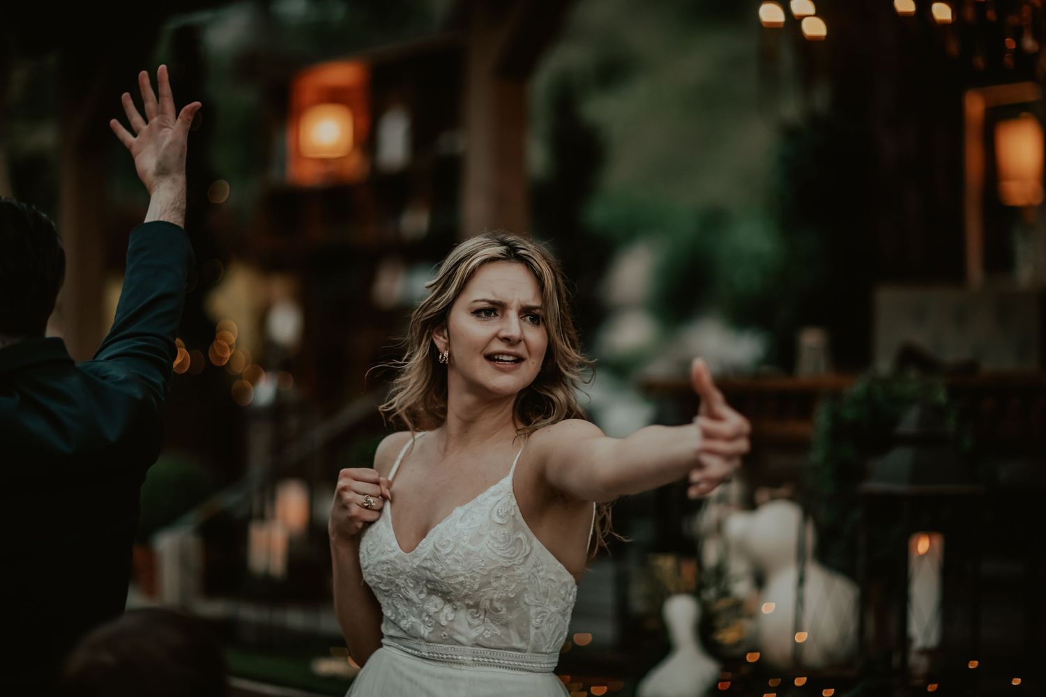 A bride in a white lace wedding dress dances with outstretched arms during an evening outdoor reception.