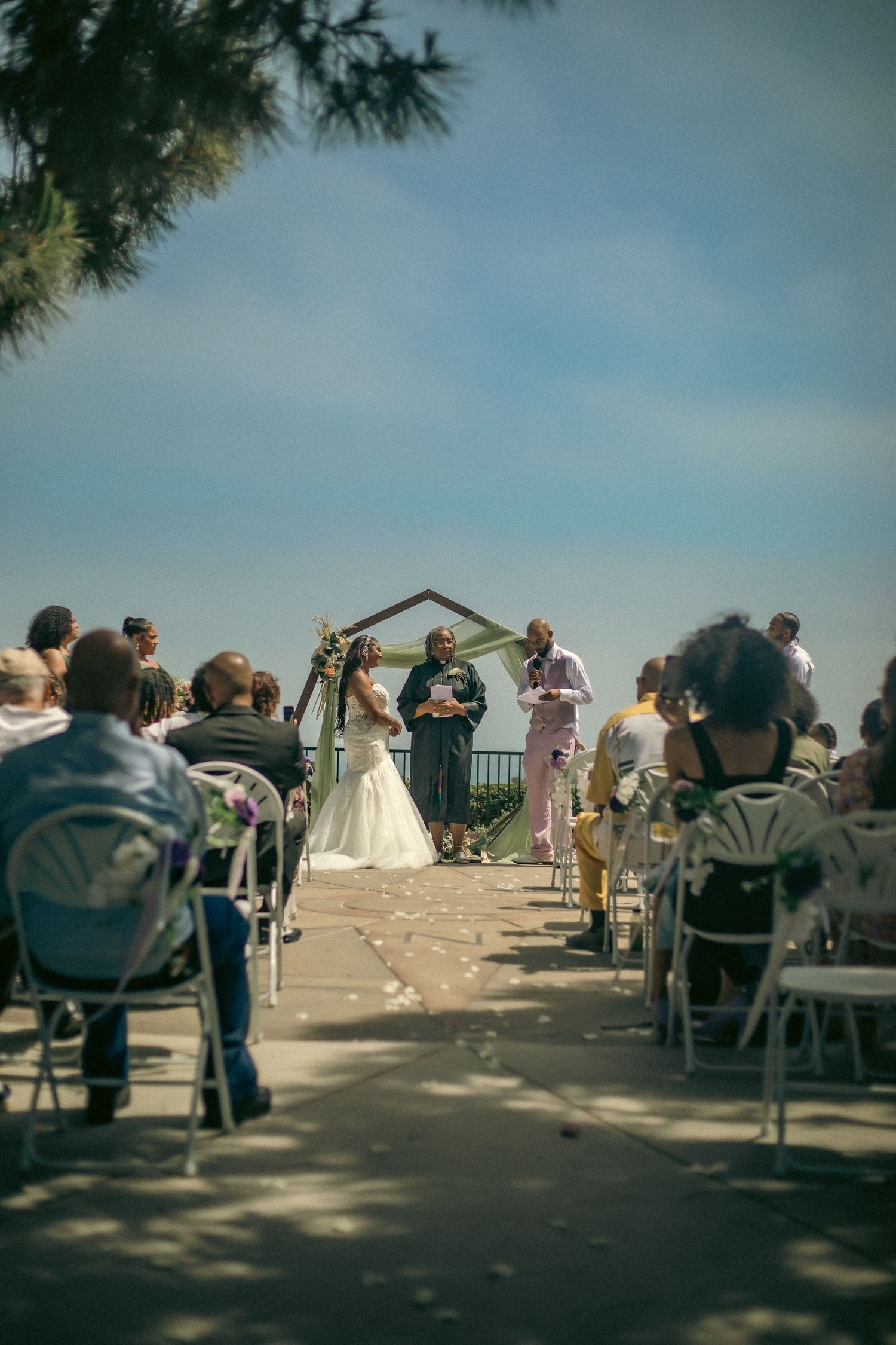 An outdoor wedding ceremony takes place under a wooden arch with guests seated in white chairs by the west coast.
