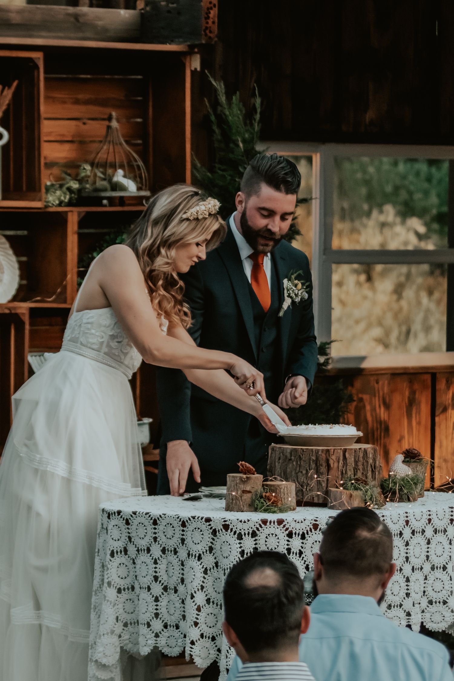 A couple cutting their rustic wedding cake together on a lace-covered table at a wooden barn venue.